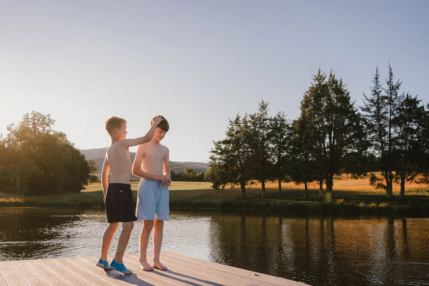 Two brothers stand on a lakeside dock at sunset, one playfully mussing the other's hair.