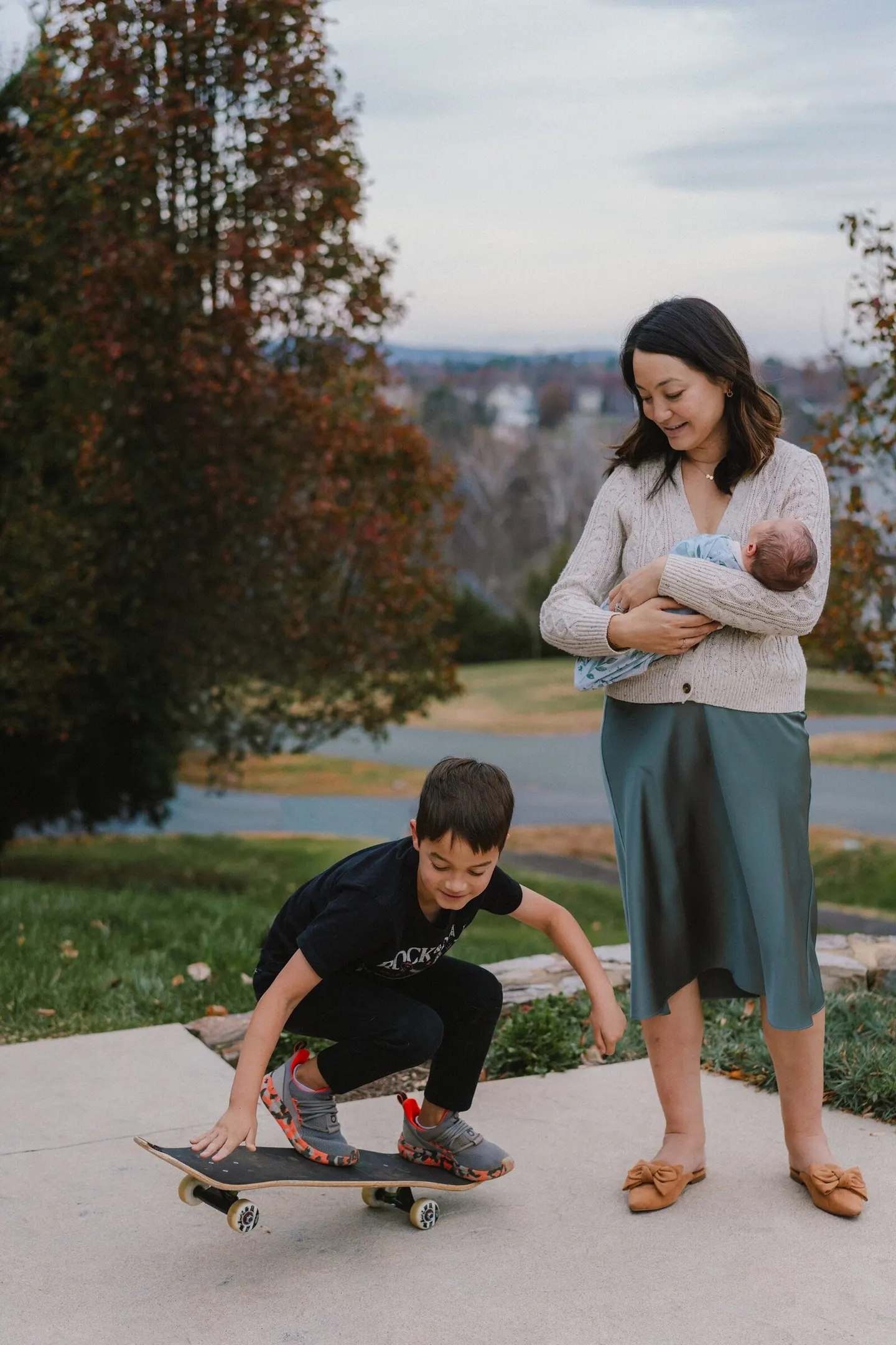 A mother holding her newborn watches her son crouch on a skateboard, hillside view behind them.
