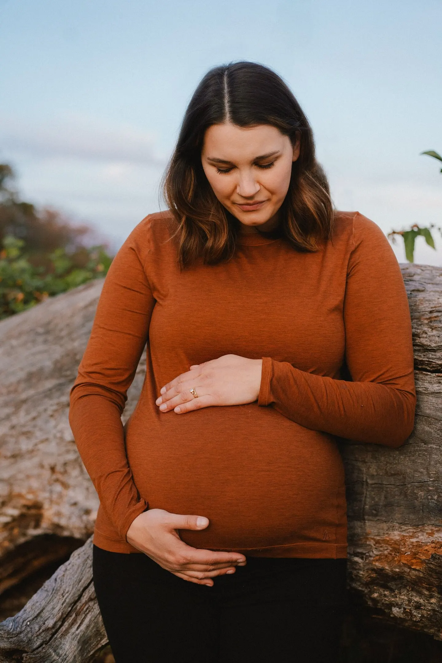 Maternity portrait in rust-colored sweater against a driftwood log at dusk