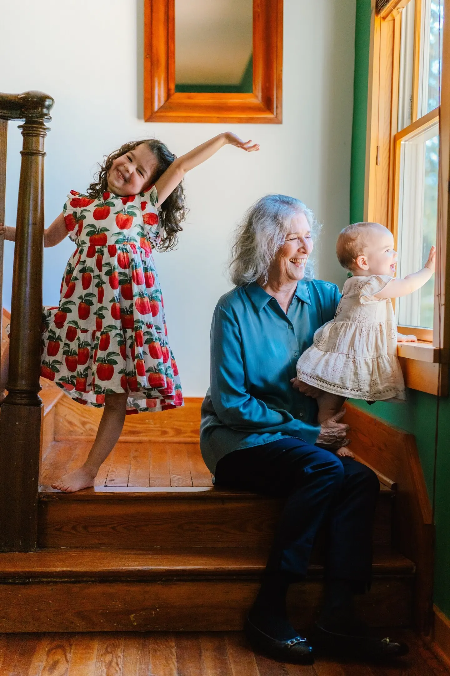 A grandmother laughs with a baby on her lap by a sunny window as an older sibling leaps nearby.