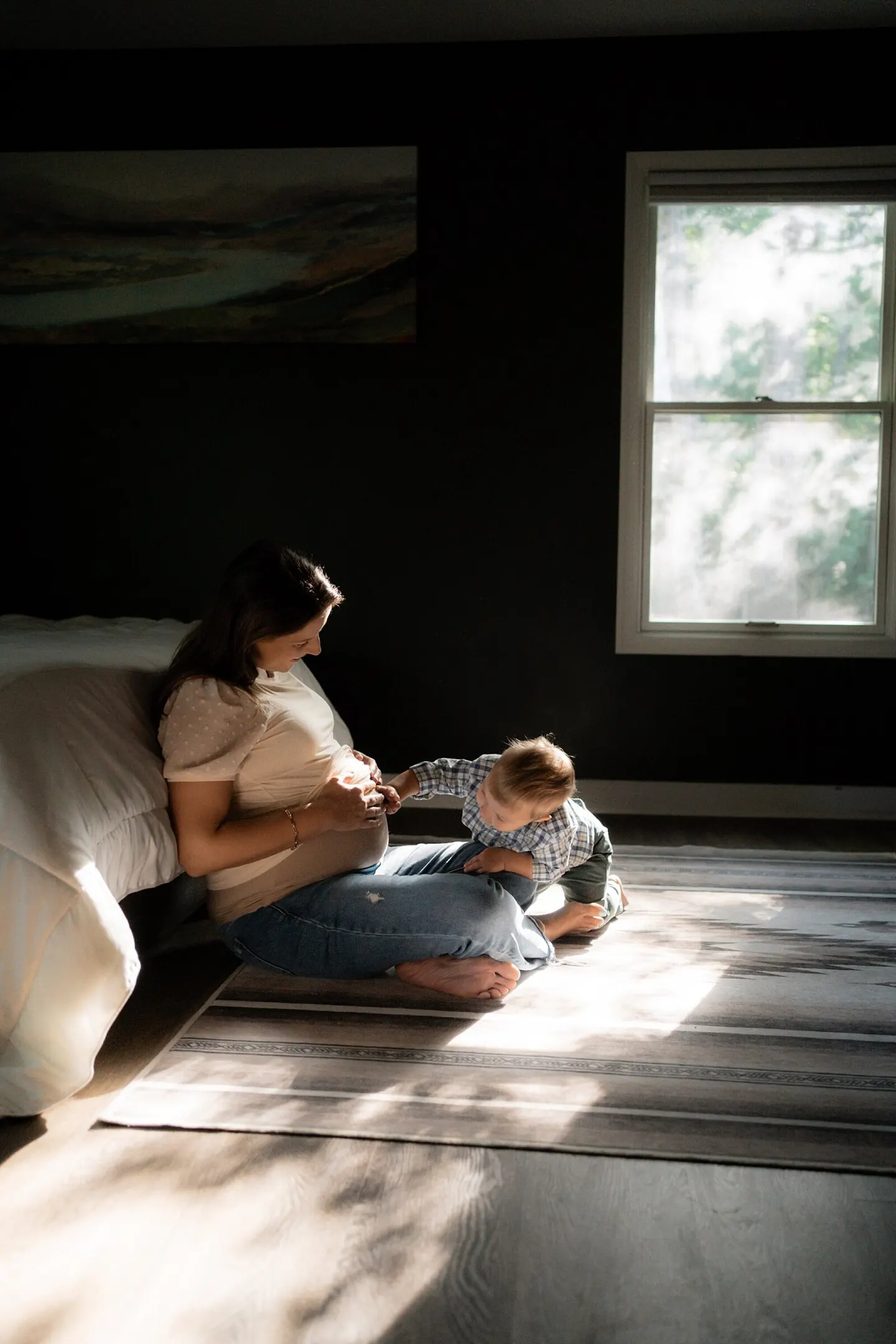 Toddler reaches for his pregnant mother's belly in a shaft of window light