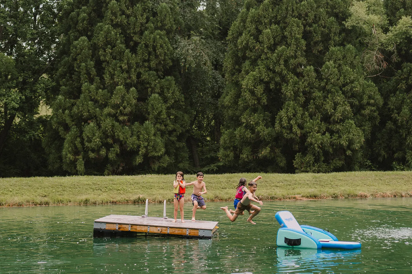 Four kids leap together off a dock into a green pond beside a floating water slide.