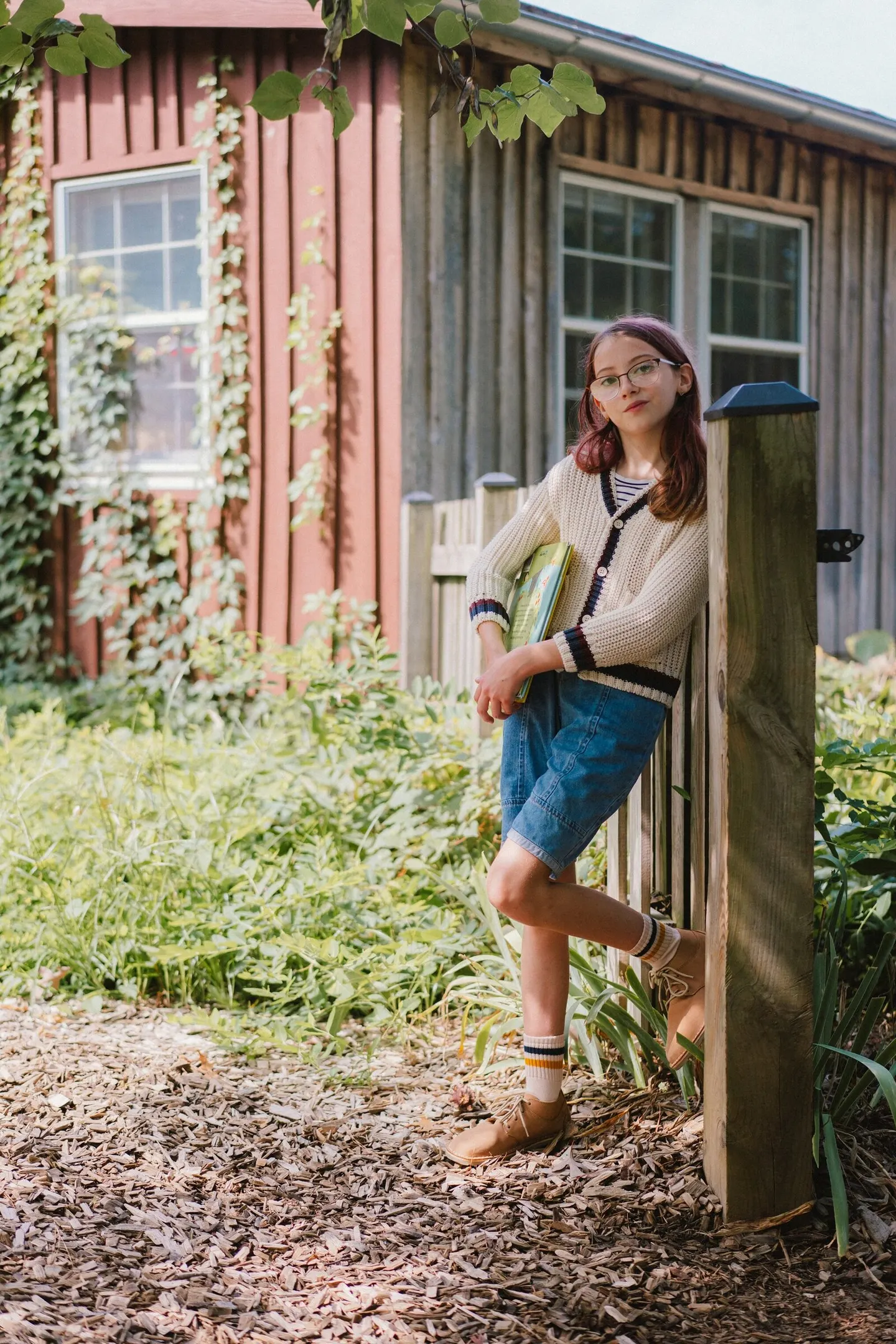 Tween girl in a knit cardigan leans against a garden fence holding a book, Charlottesville portrait