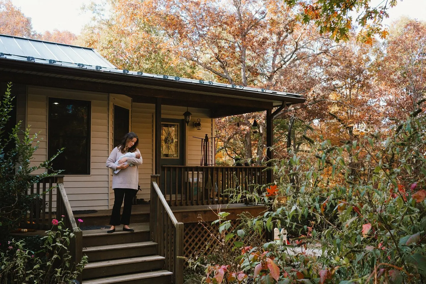Mother carries her newborn up the porch steps of a fall cabin surrounded by turning leaves
