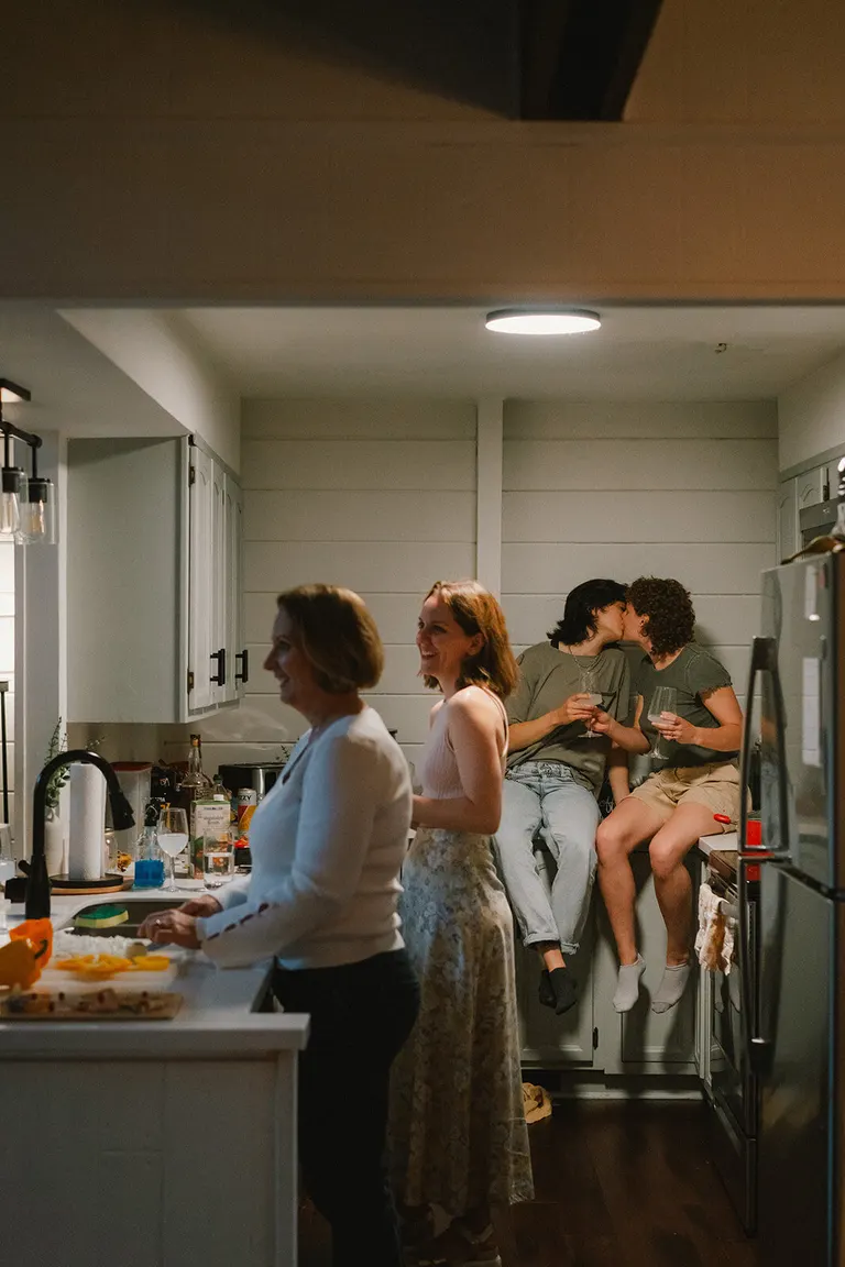 A young couple shares a kiss on the kitchen counter while relatives chat and cook nearby.