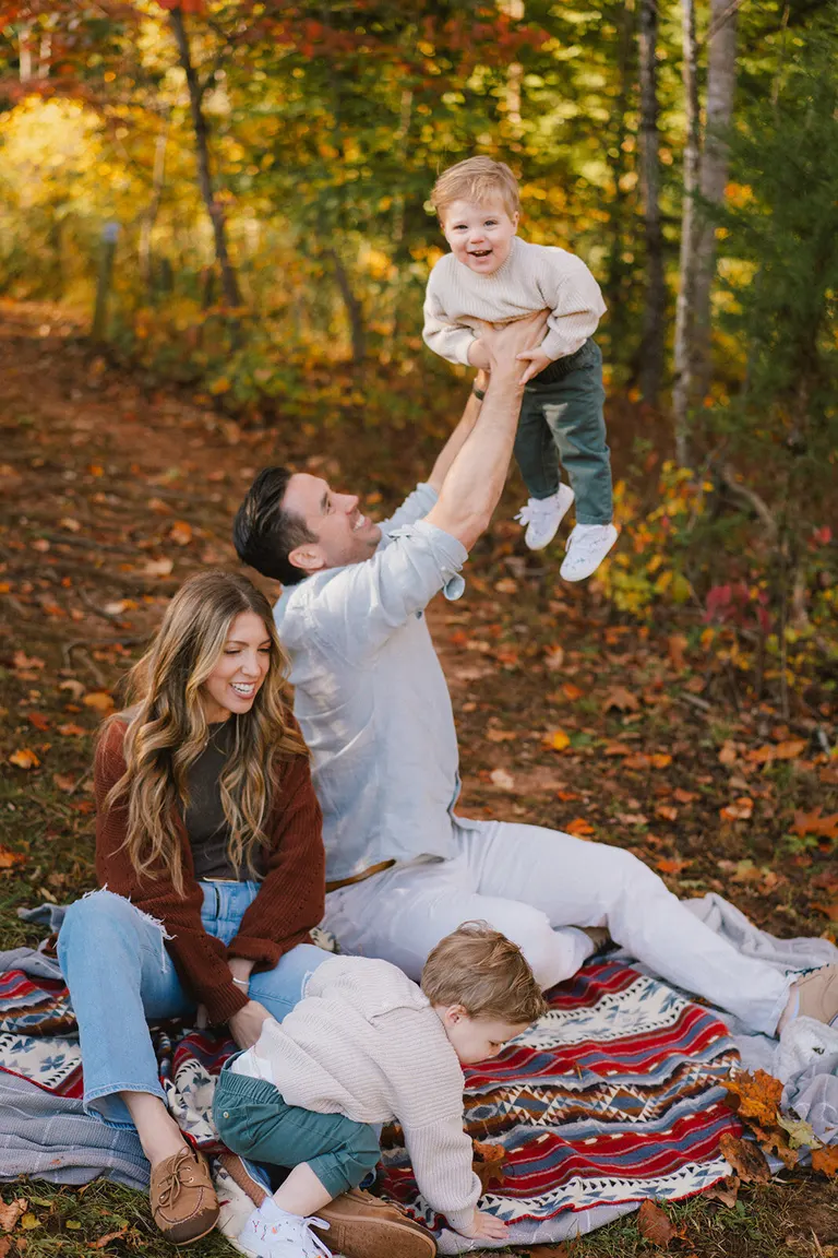 A father lifts his toddler high overhead in the autumn woods while mom and a sibling laugh nearby.