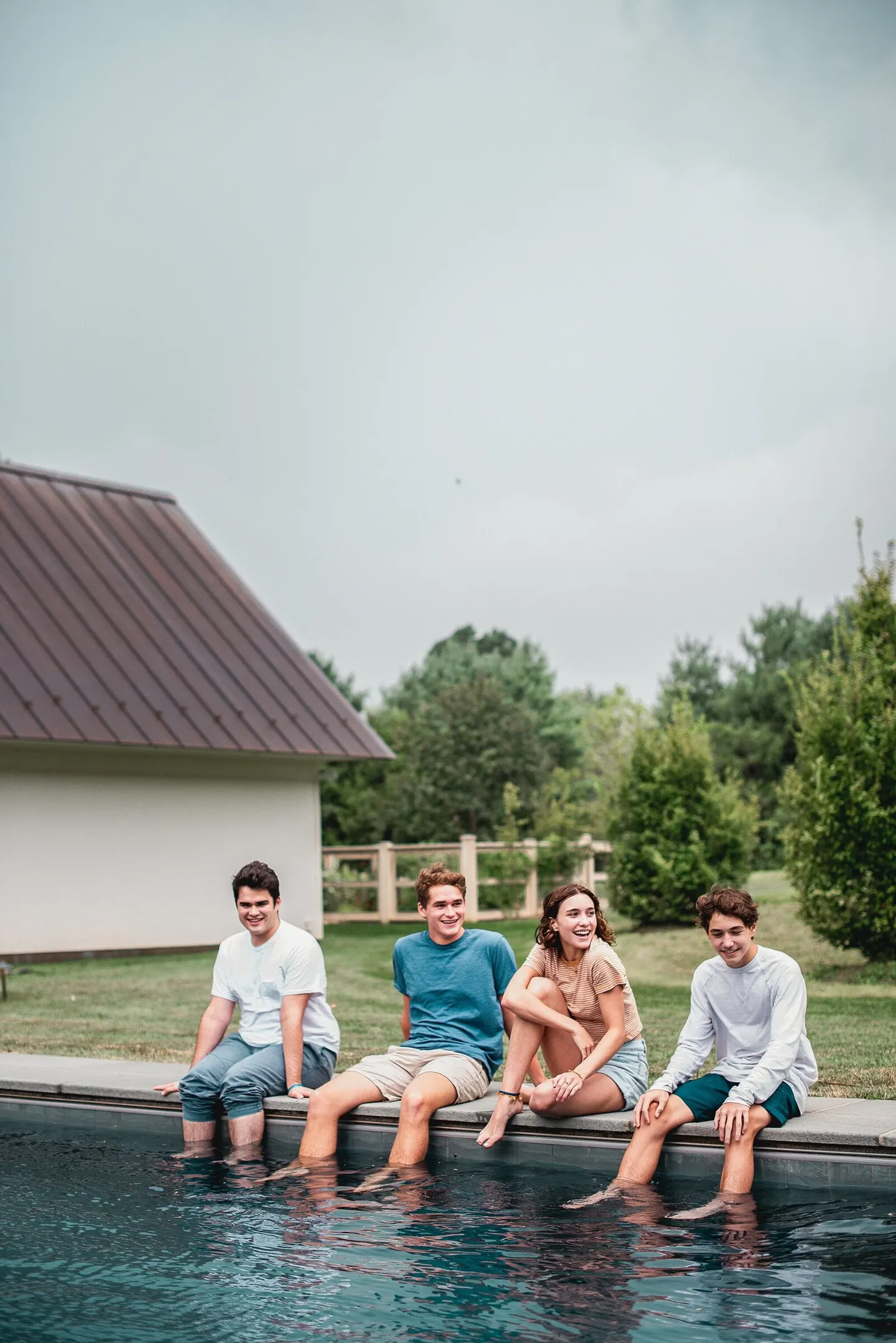 Four cousins sit laughing along a pool's edge with their feet in the water under a cloudy sky.