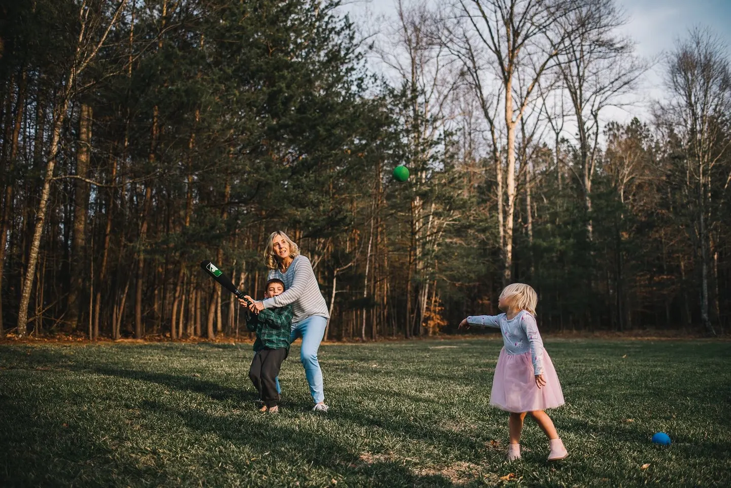 A grandmother helps her grandson swing a bat while a little girl tosses him a ball across the backyard grass.