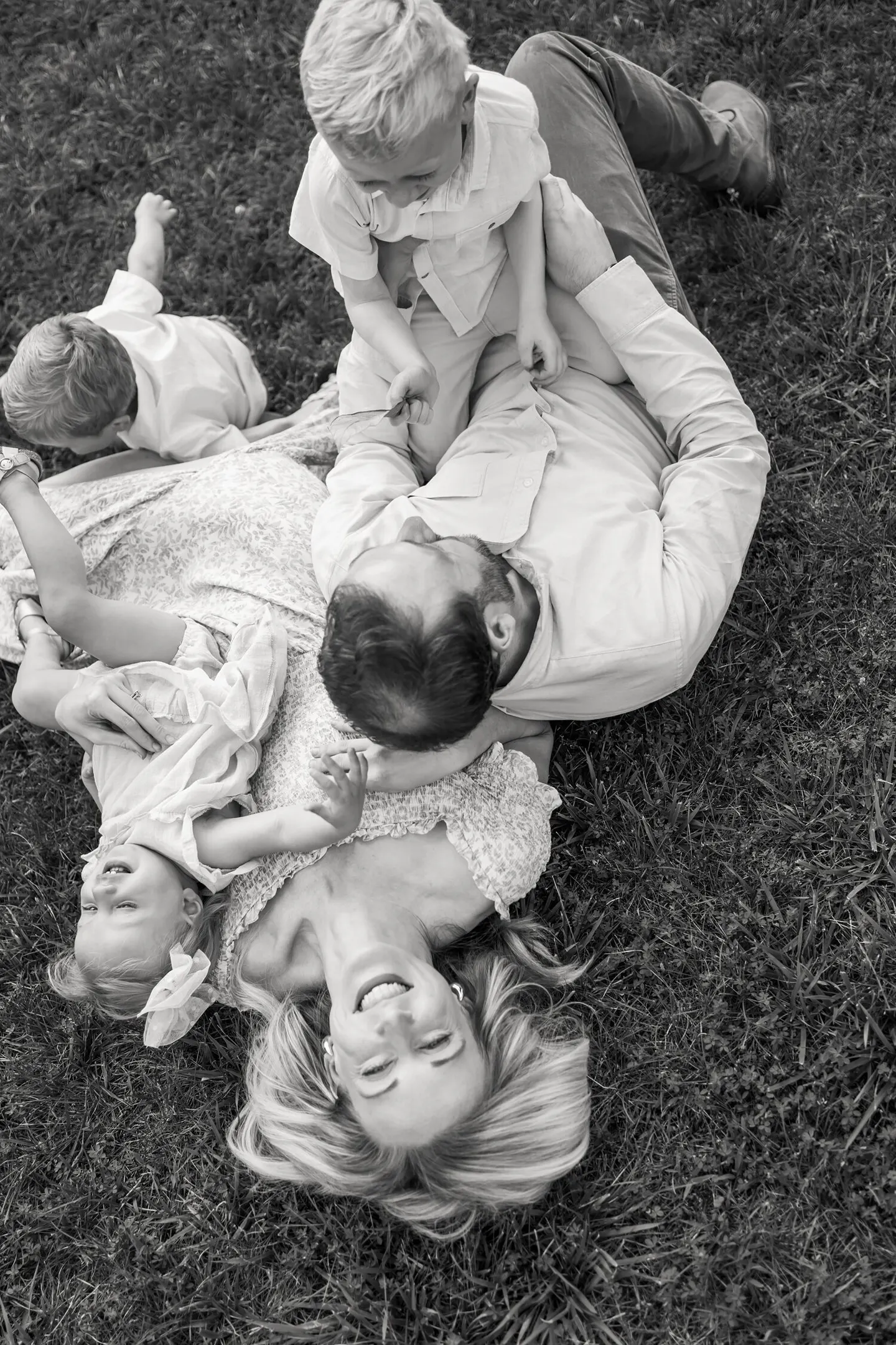 A family piles together laughing in the grass, kids climbing over their parents in playful chaos.
