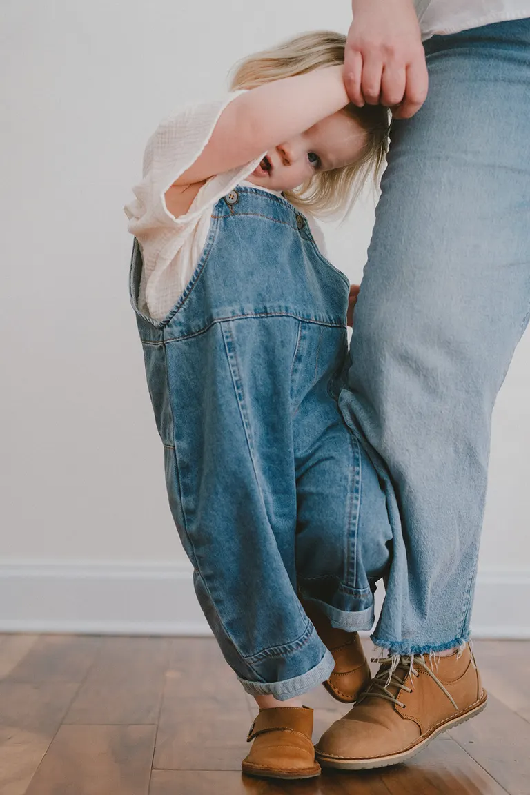 Toddler in denim overalls peeks out shyly while gripping a parent's arm indoors