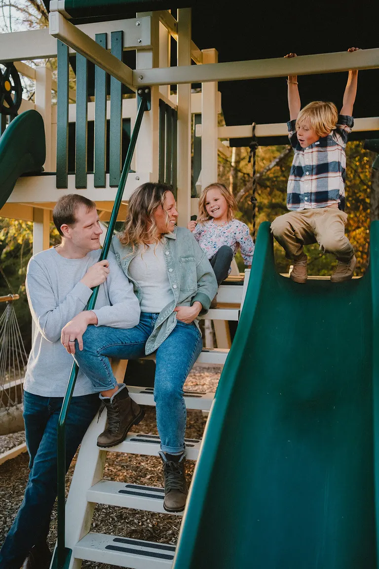 Parents sit together on a backyard play-structure while their kids climb, slide, and hang from the monkey bars.
