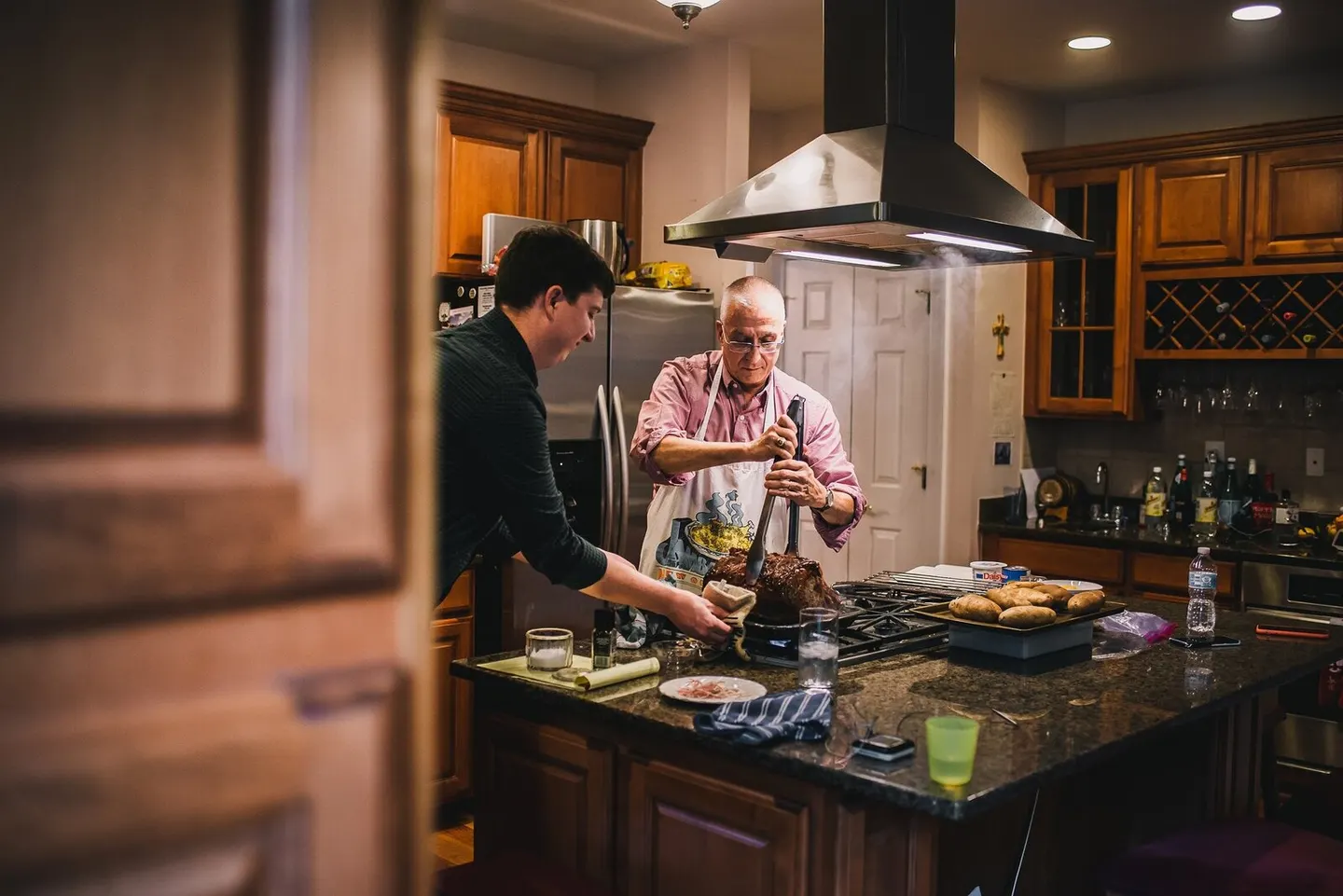 A grandfather in an apron carves a roast at the stove while his grandson holds the platter steady beside him.