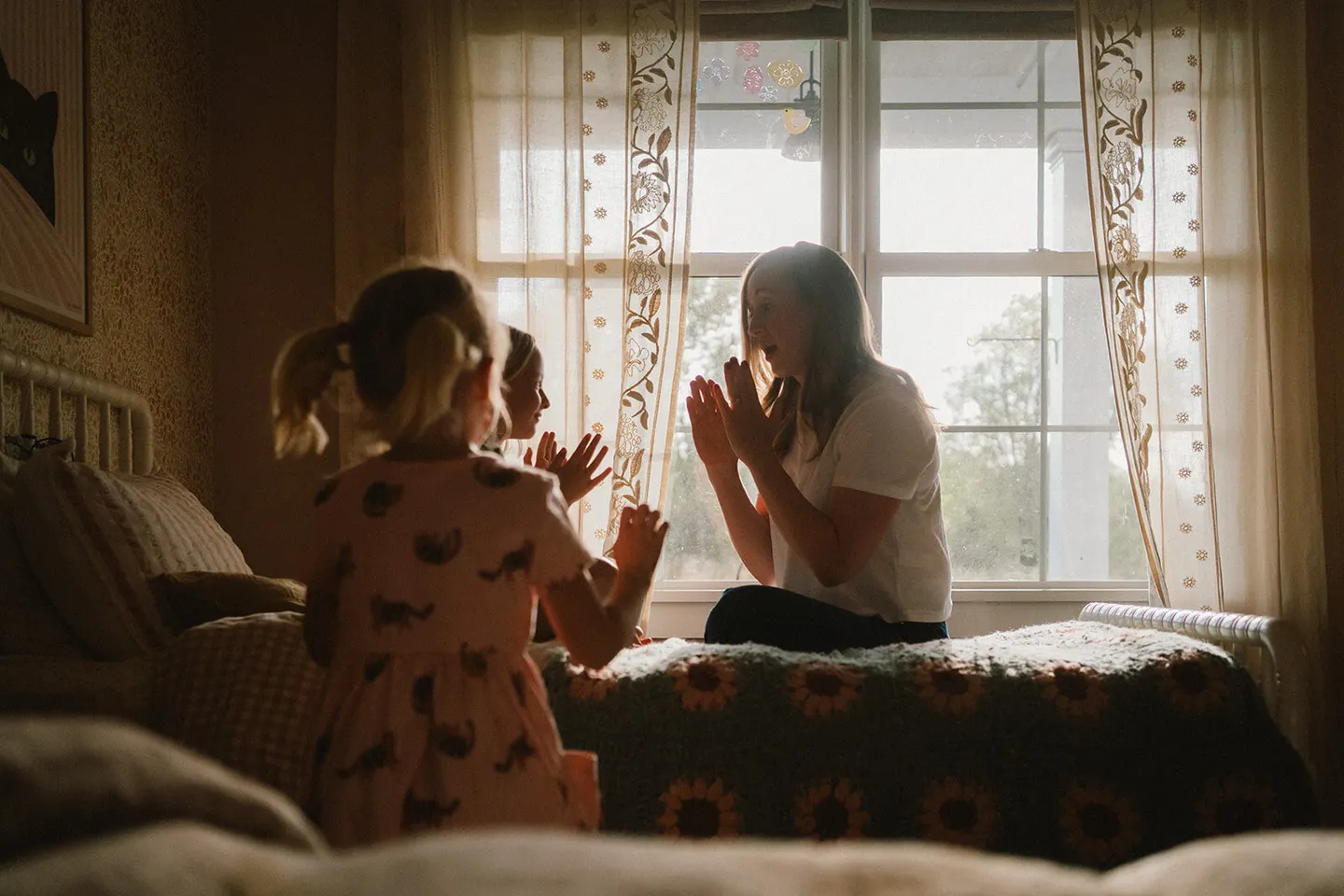 A Charlottesville family photographer catches a mom and two girls in a clapping game by a sunlit window.