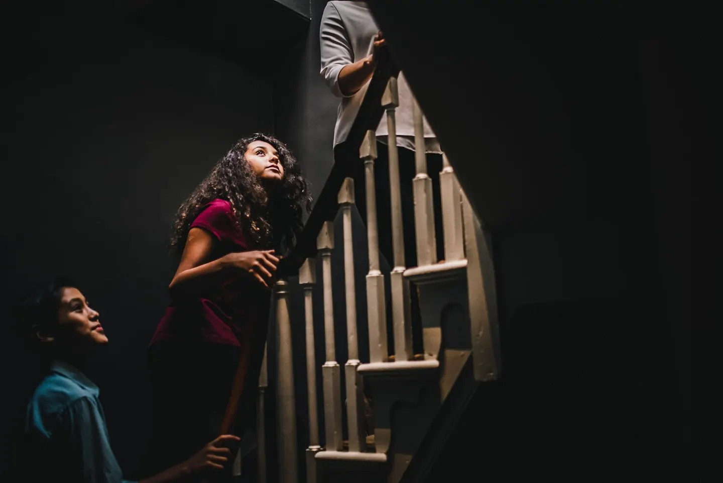 Backlit teens climb a dim staircase, one glancing upward as light spills from above