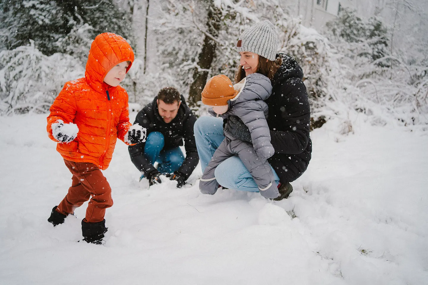 A family snowball fight breaks out in falling snow, a baby bundled up watching from mom's arms.