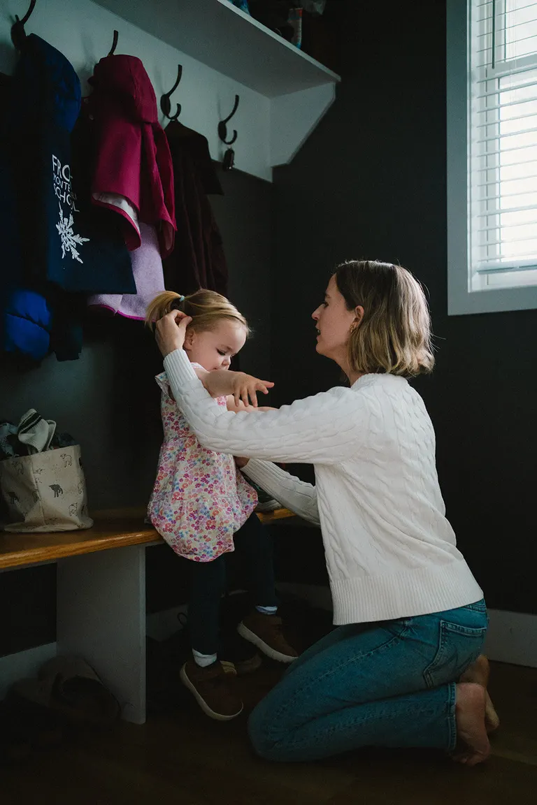 In a cozy mudroom, a mother fixes her daughter's ponytail beneath a row of coat hooks.