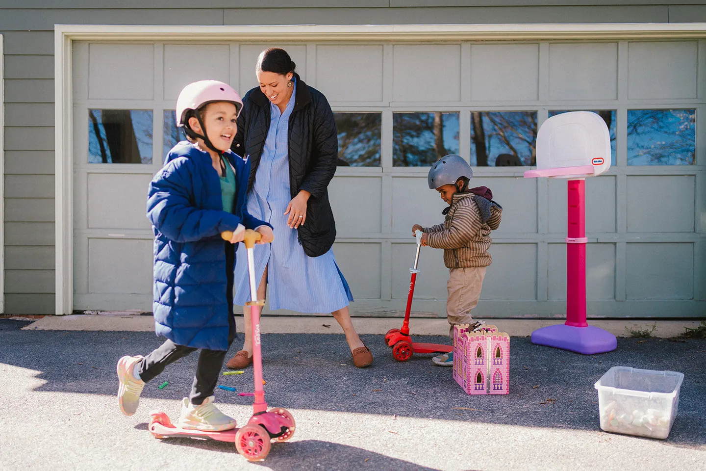 A mom watches her helmeted kids race scooters down the driveway past the garage.