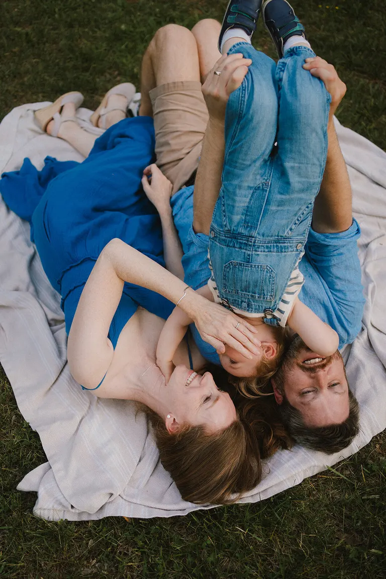 Parents lie head to head on a blanket, laughing as their toddler daughter peeks between them.