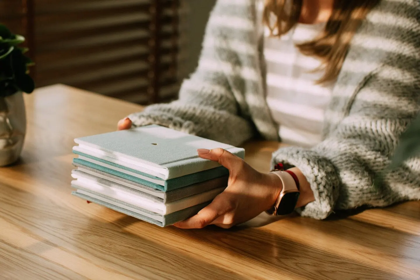 Stack of small square albums in sage and cream cradled in two hands at a sunlit table