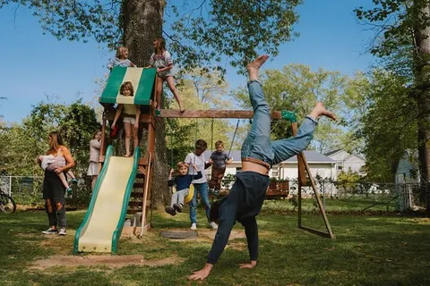 An adult cartwheels through the yard while cousins climb a wooden playset and a mother holds her baby.