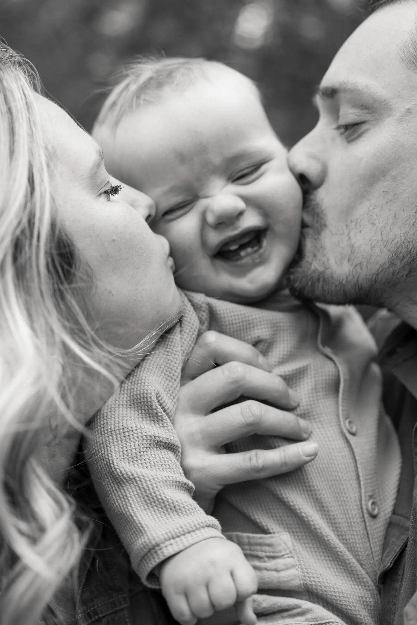 A close black-and-white portrait shows two parents kissing their giggling baby on both cheeks.