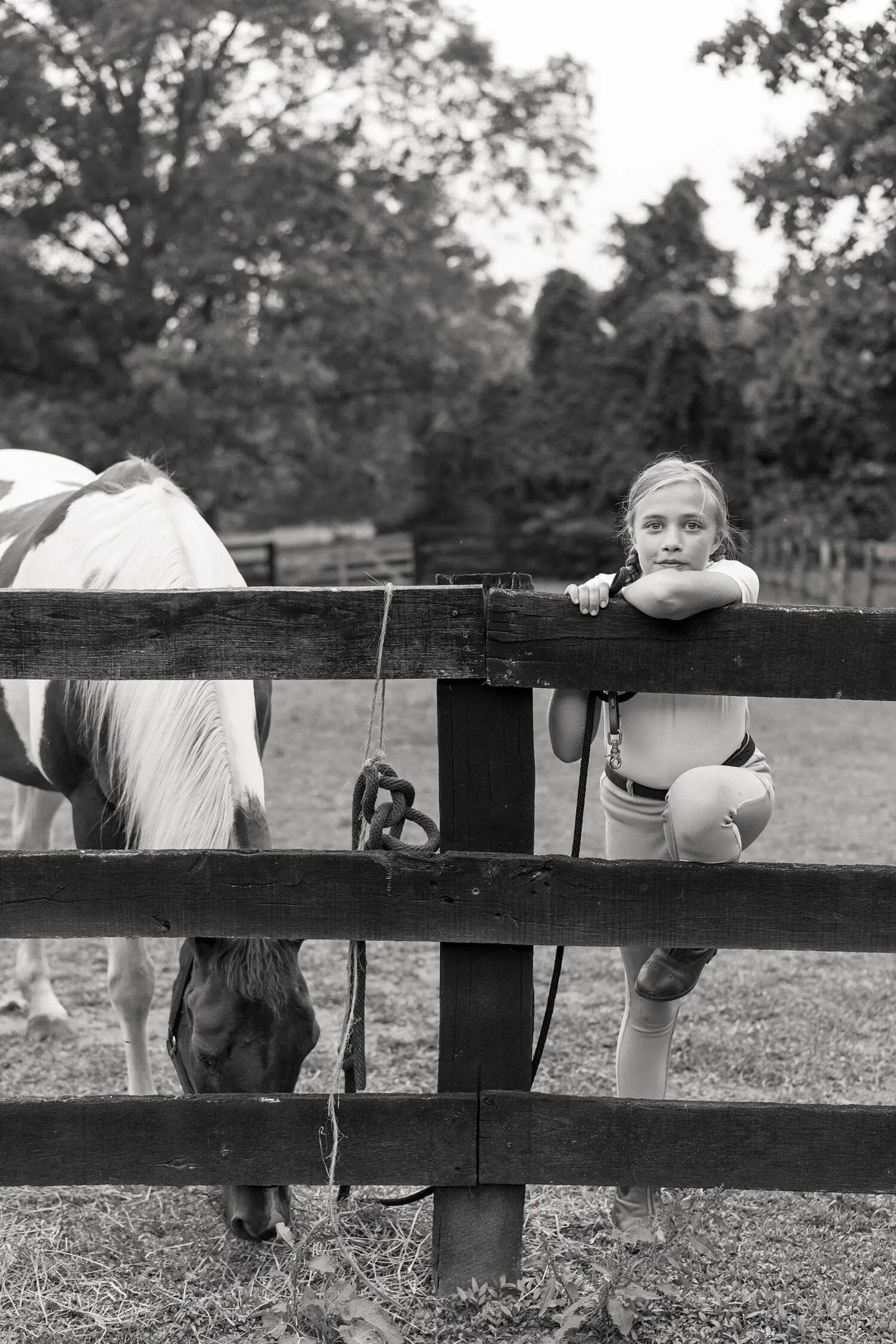 In black and white, a young rider in a helmet leans toward a pony at the fence line.