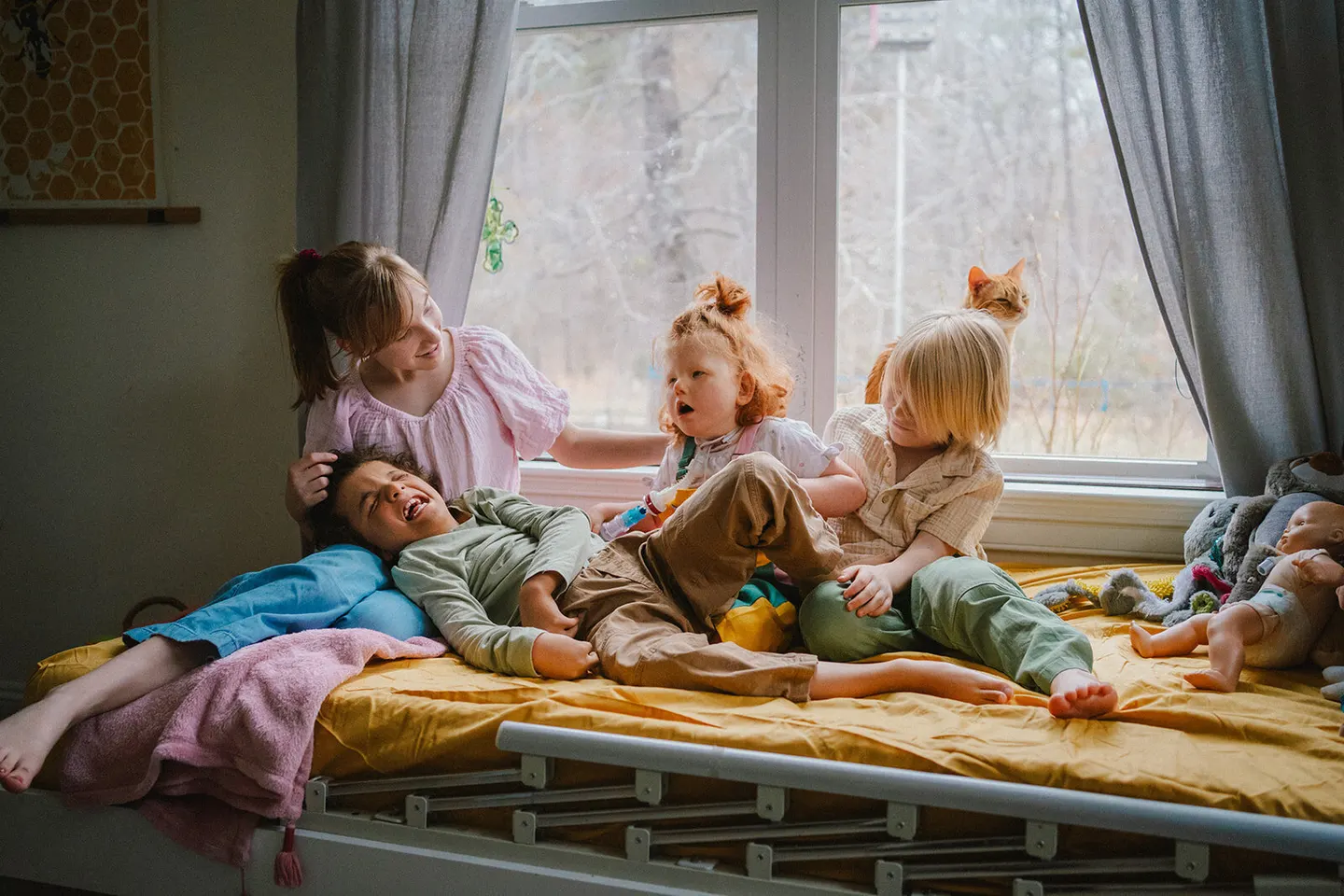 Two kids pile onto a sunlit bed with the family cat, framed by a bright window.