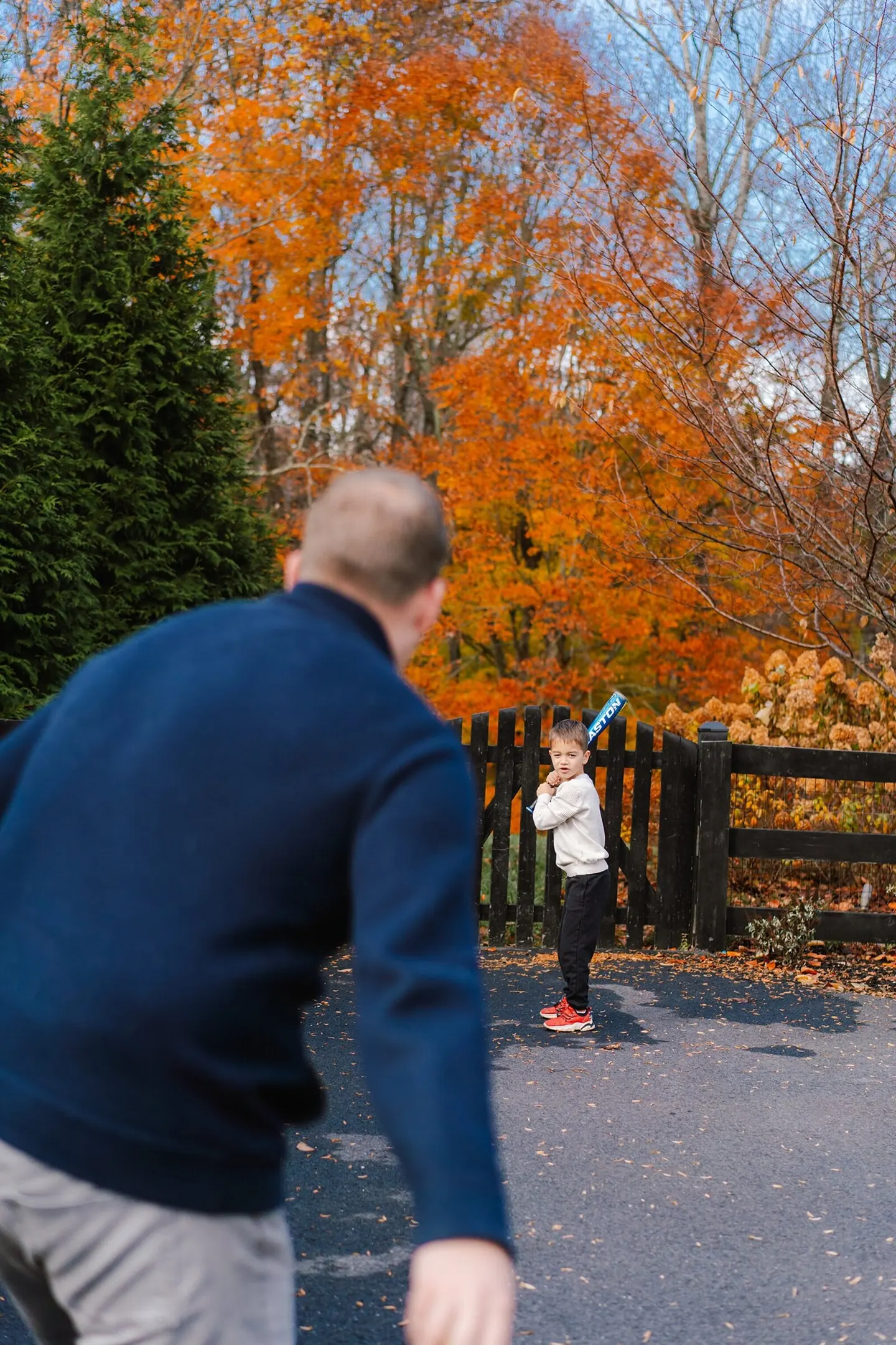 A blurred father watches his son take a batting stance in the driveway under blazing orange fall trees.