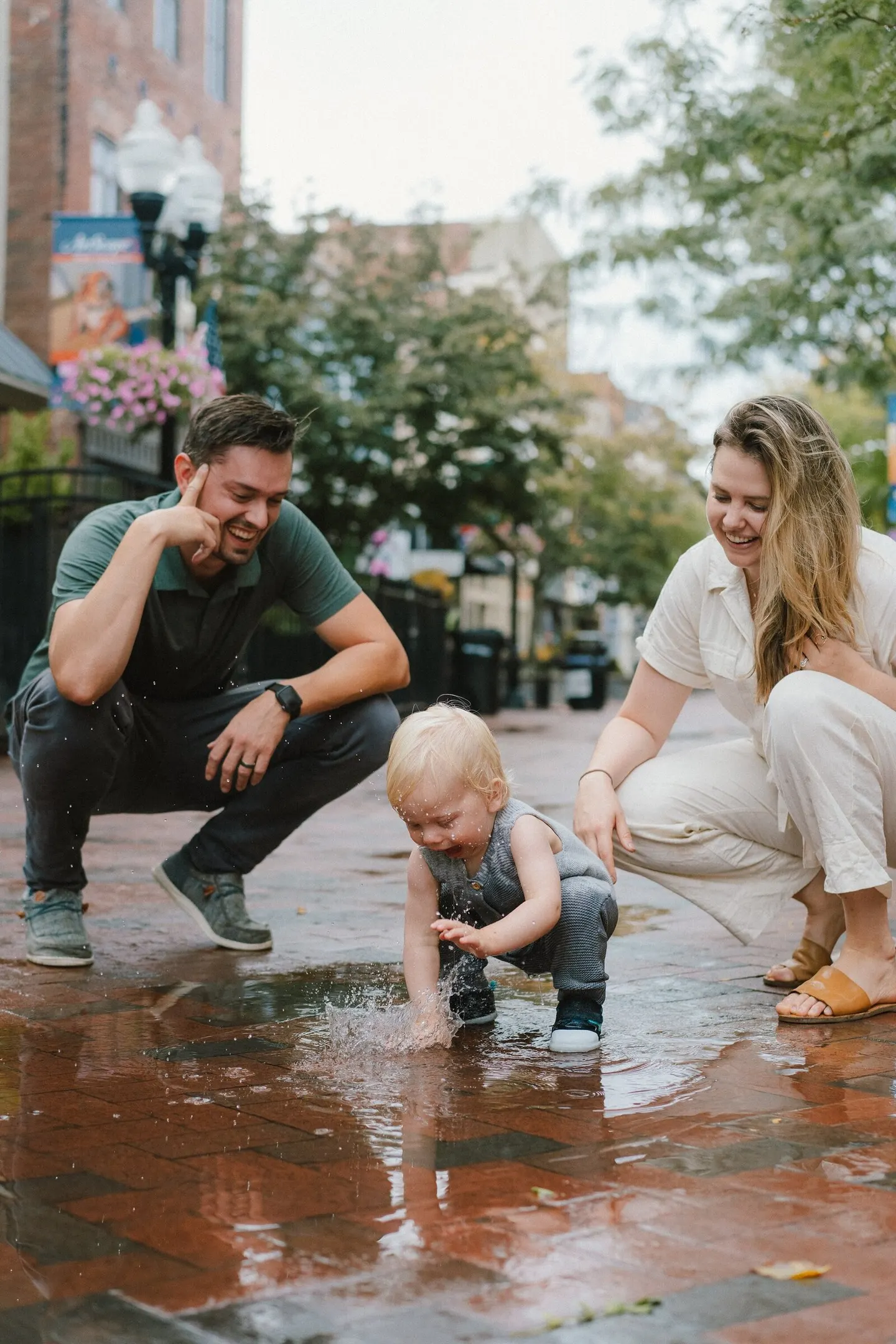 A toddler stomps through a rain puddle downtown while his laughing parents crouch beside him.