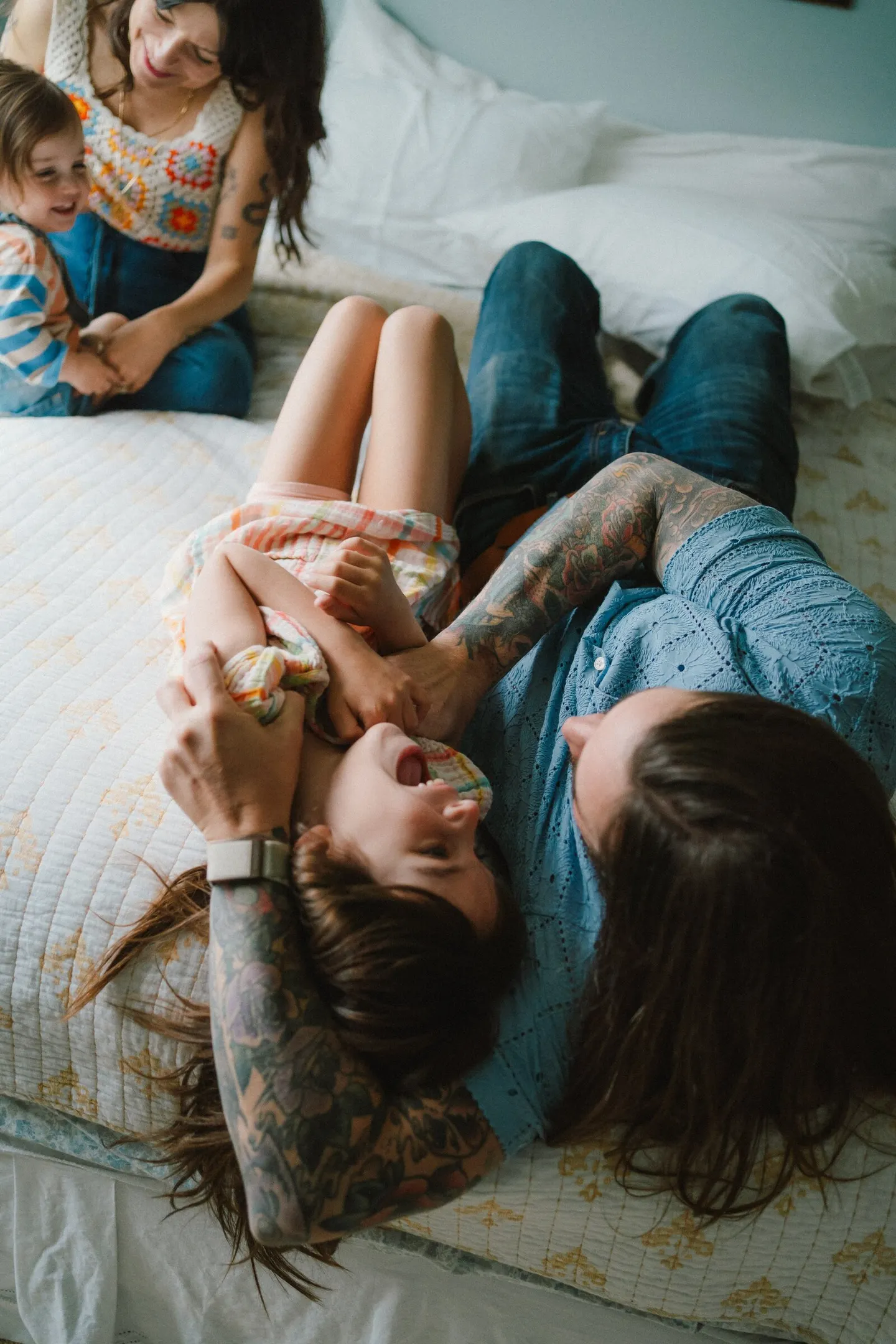 Seen from above on a bed, a dad tickles his daughter while mom cuddles a toddler close by.