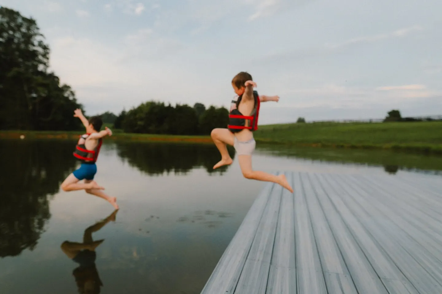 Two boys in life vests leap off a wooden dock into a green pond mid-summer.