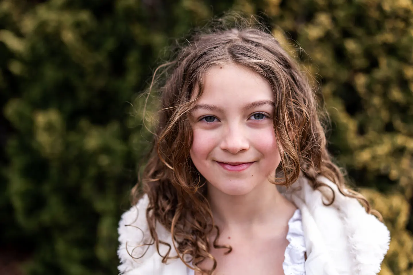 Girl in a white knit wrap smiling in front of evergreen hedges