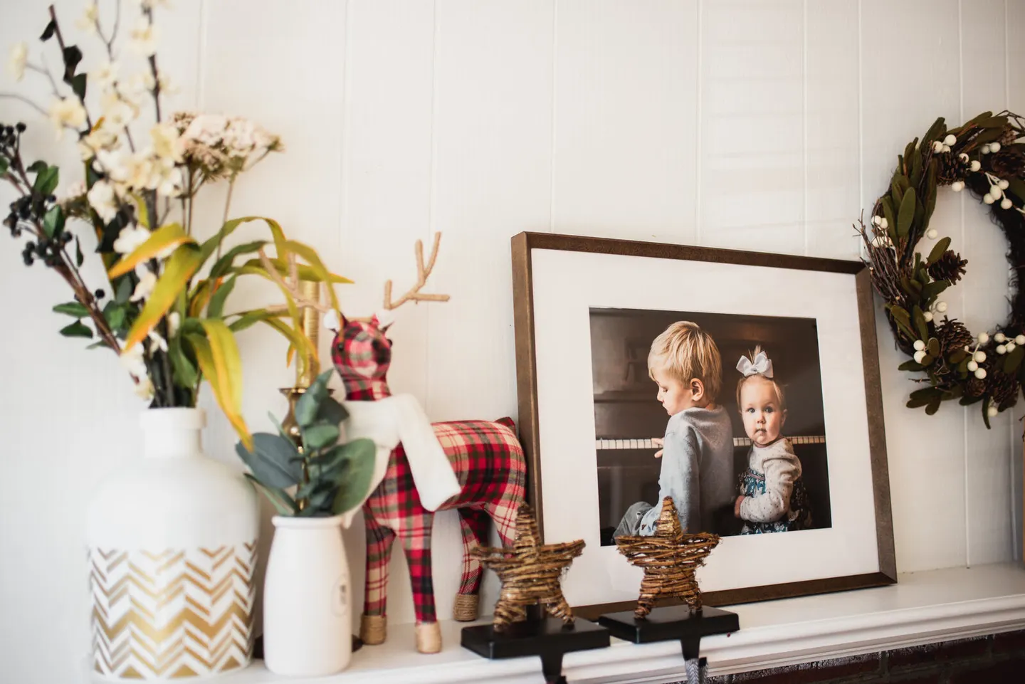 Piano-bench siblings framed in bronze on a holiday mantel beside a plaid reindeer and wreath