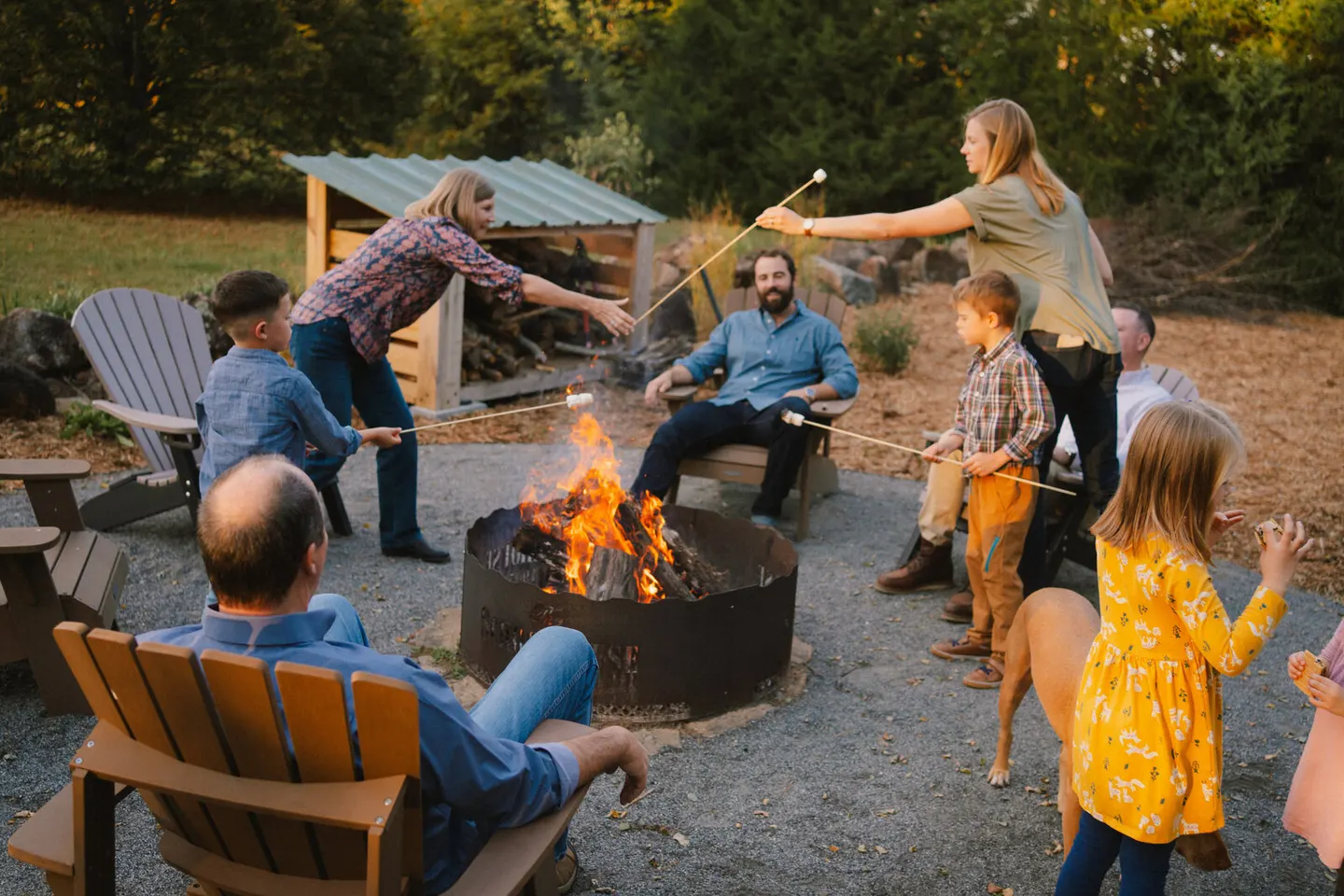 Kids and adults roast marshmallows together around a crackling backyard fire pit at sunset.