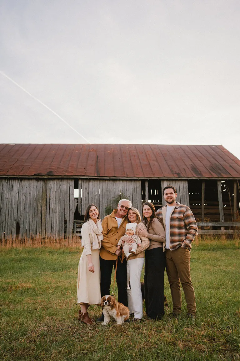 Three generations and their spaniel gather in front of a weathered barn, the baby held close in a knit cap.