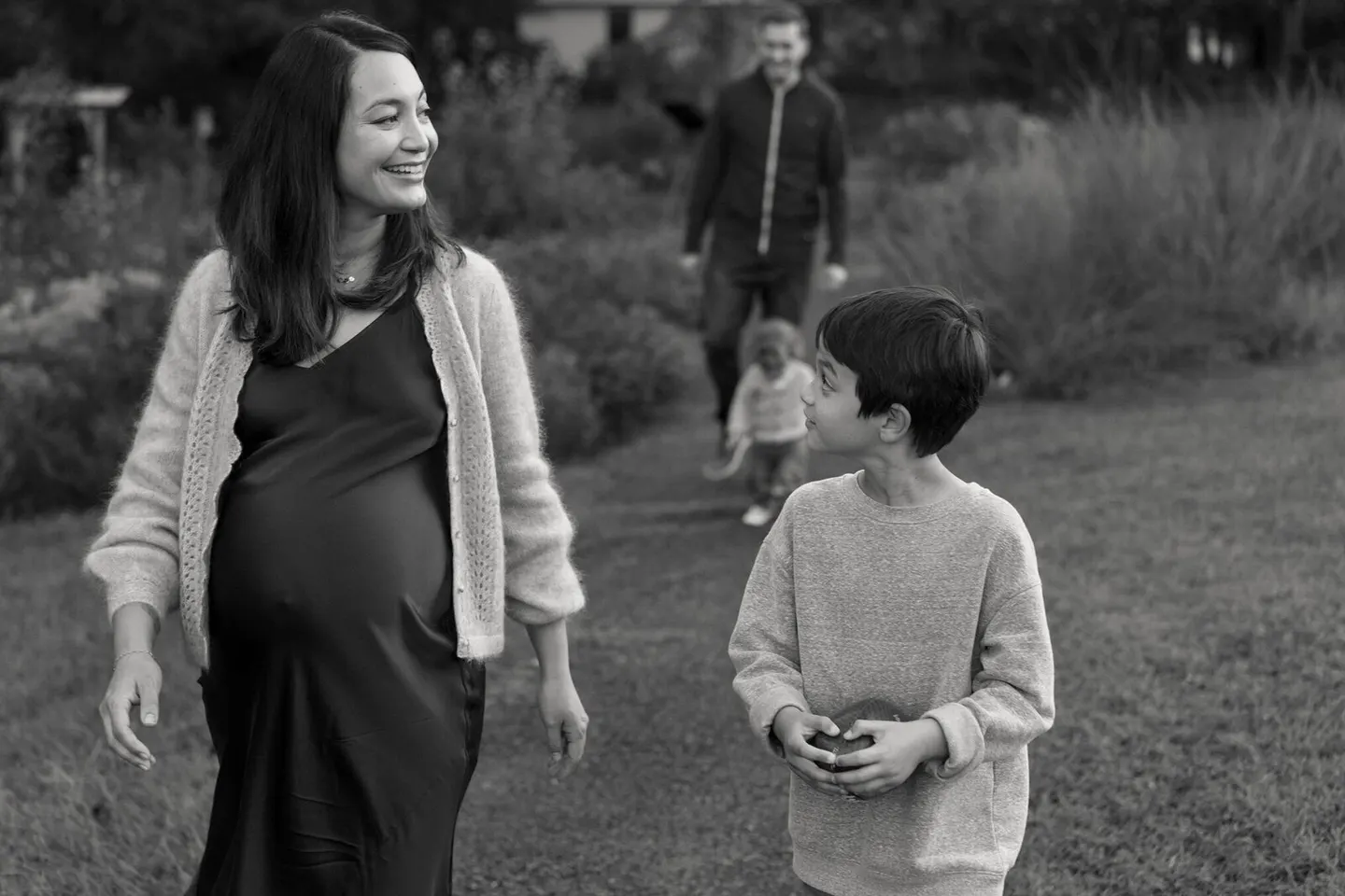 Pregnant mother walks a garden path smiling at her son while dad and toddler trail behind