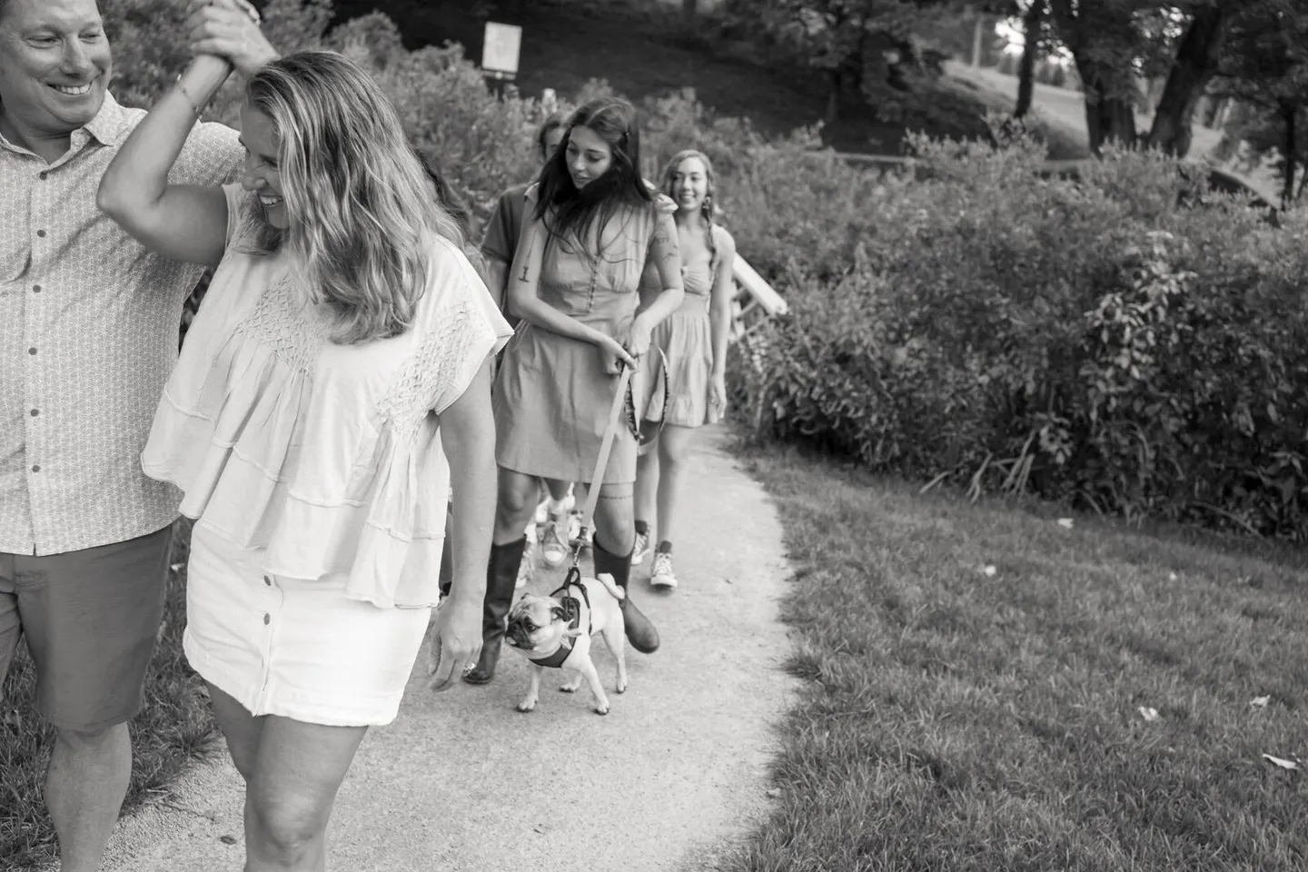 In black and white, a multigenerational family strolls a garden path with their pug on a leash.