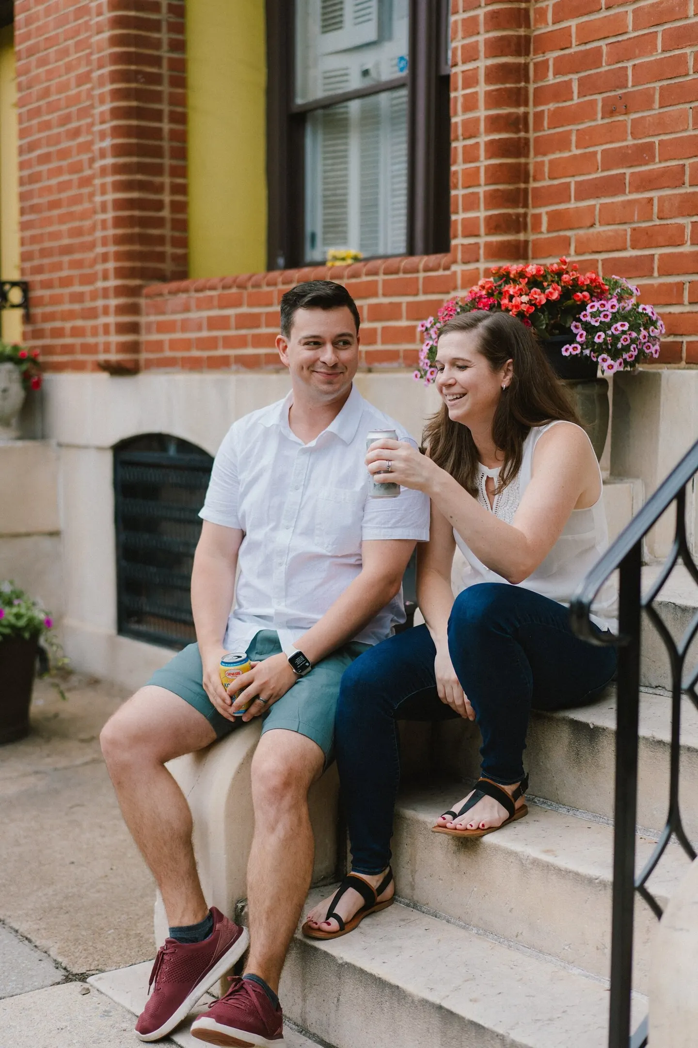 A couple relaxes on brick porch steps with drinks in hand, surrounded by blooming flowers.