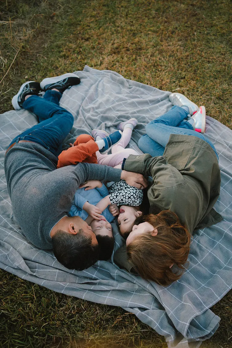 Parents cradle two giggling toddlers in a tangle of arms on a plaid picnic blanket in the grass.