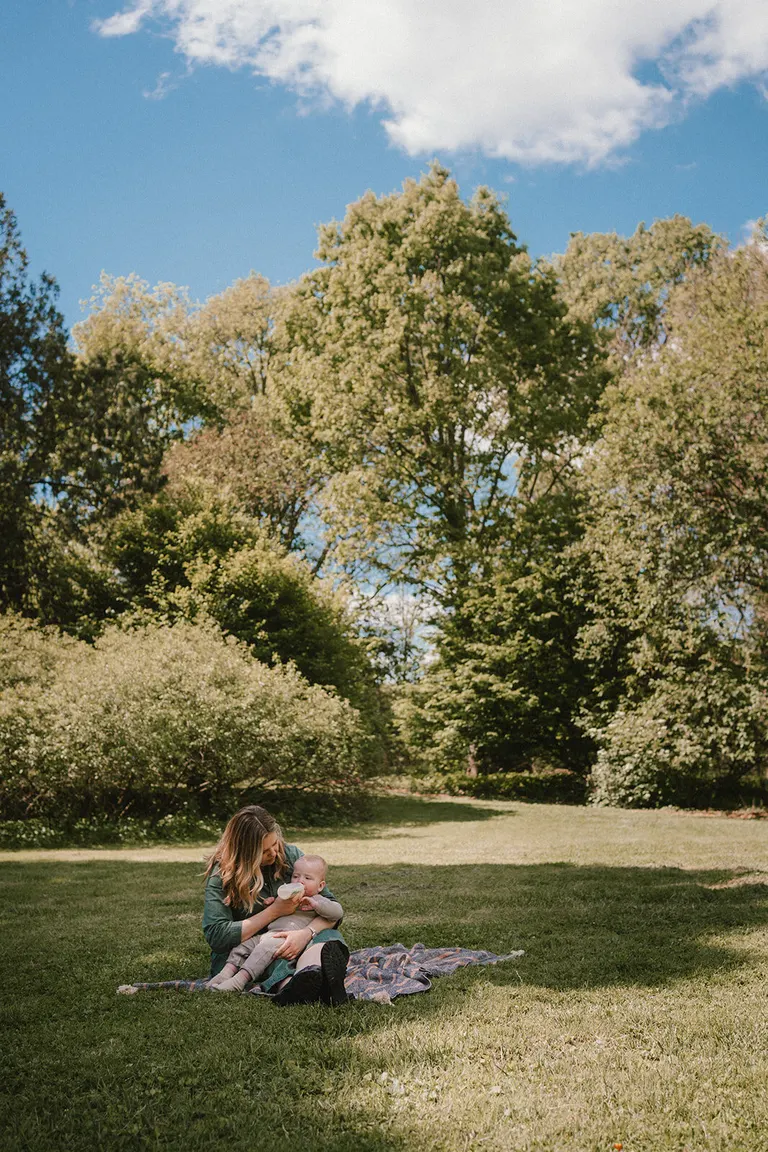A mother feeds her baby a bottle while seated on a blanket in a sunny park.