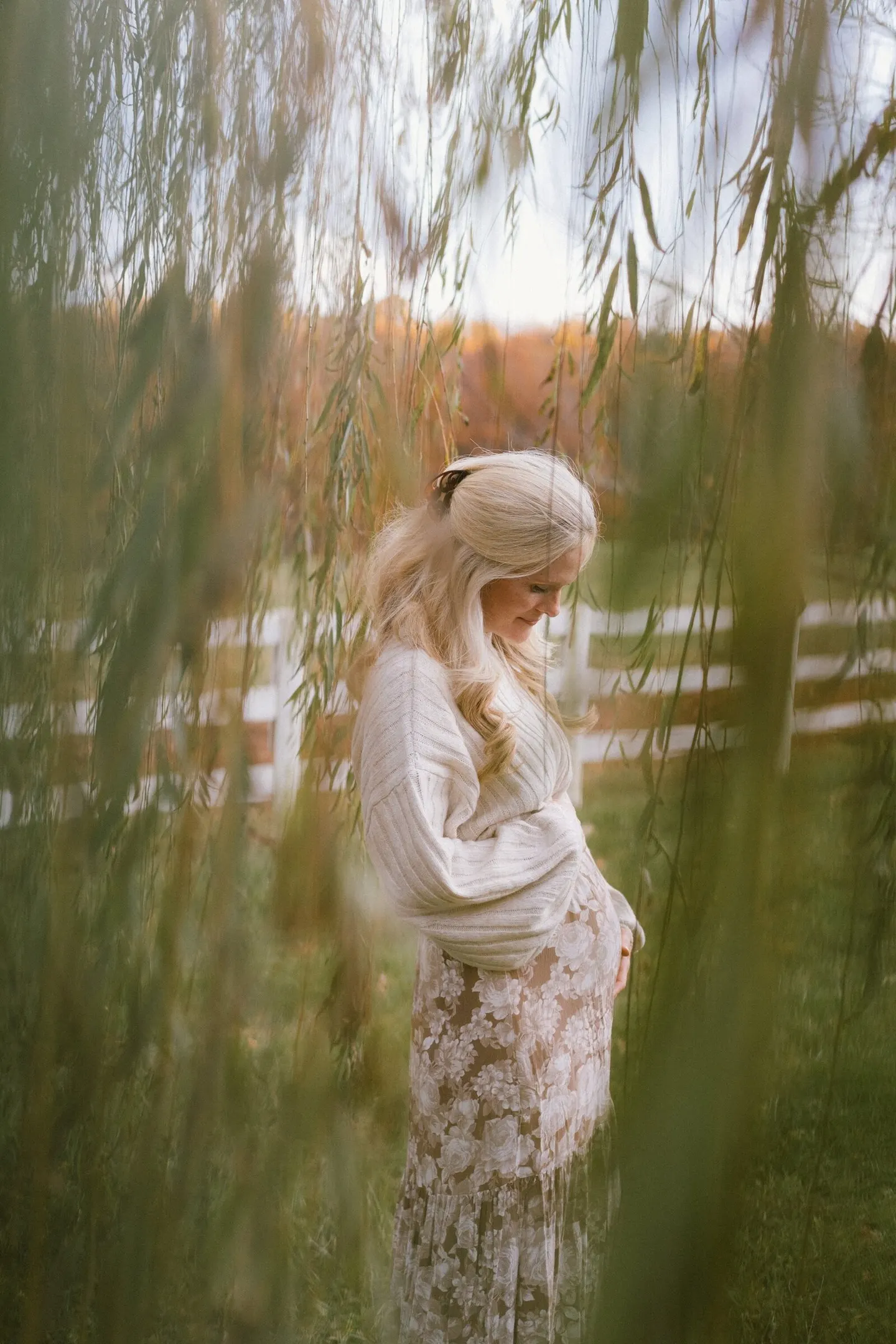 Pregnant woman in a lace skirt stands beneath willow branches near a white fence