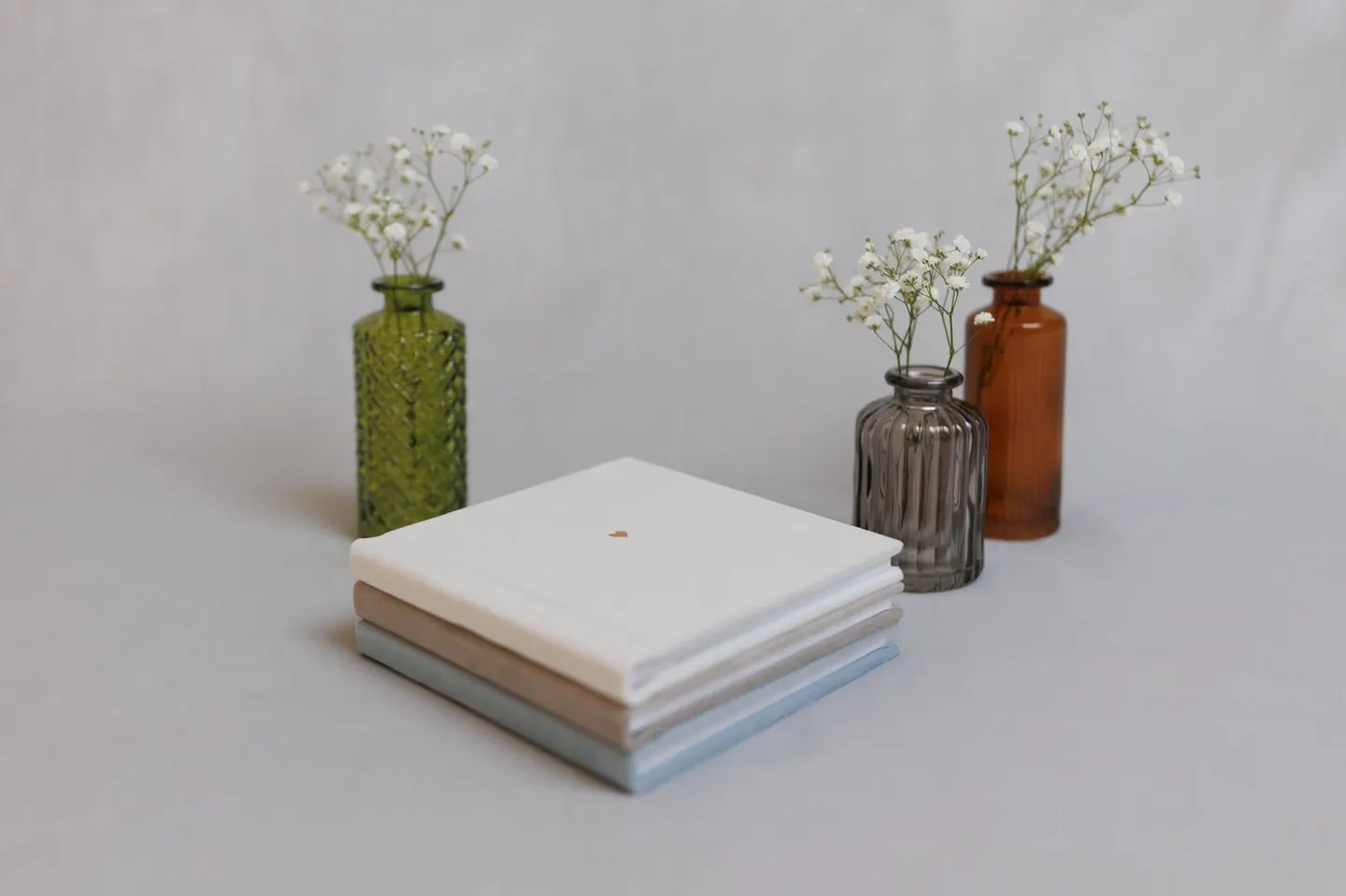 Trio of small square albums in white, taupe, and dusty blue staged beside bud vases