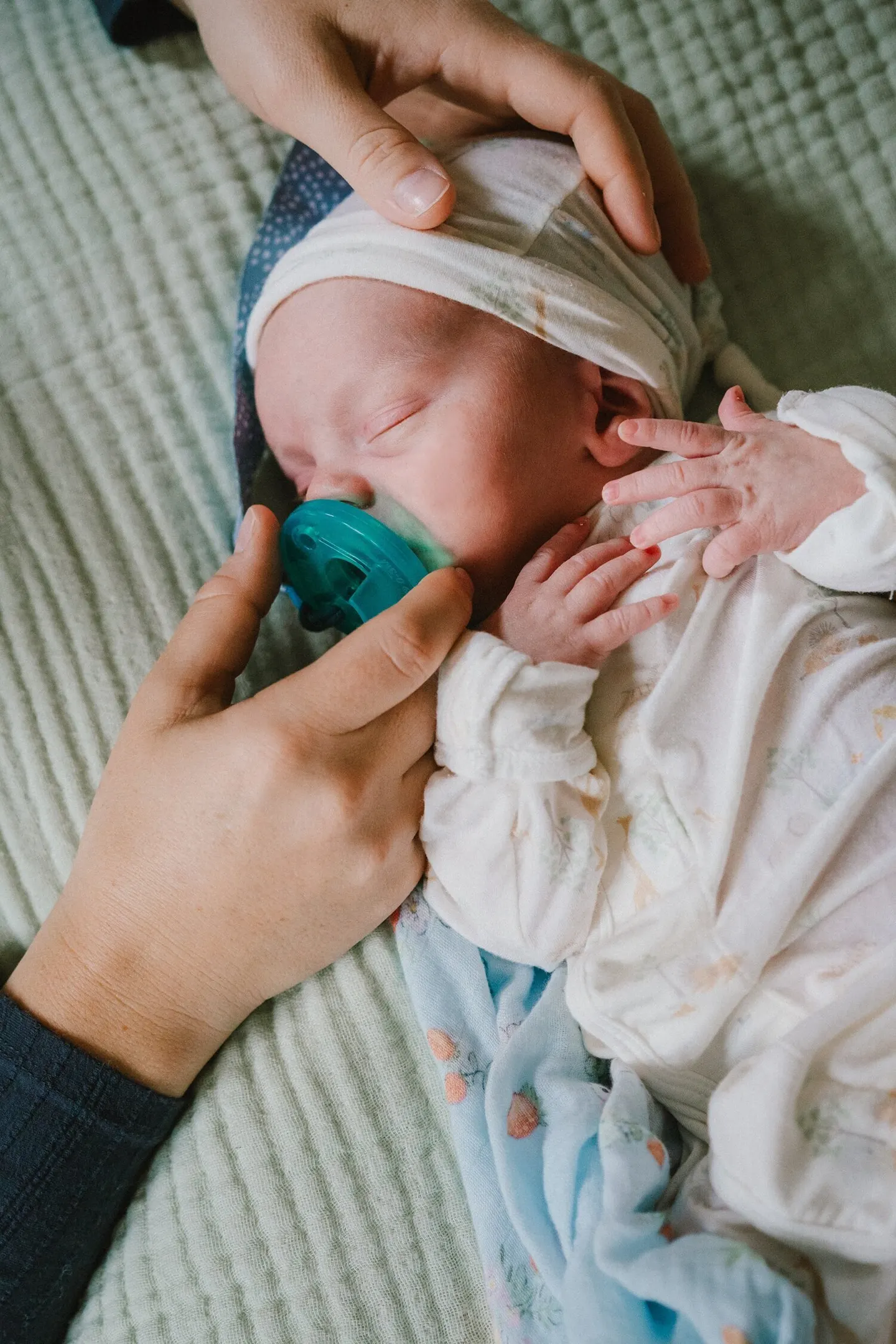 Overhead close-up of a newborn in a knotted headband gripping a pacifier