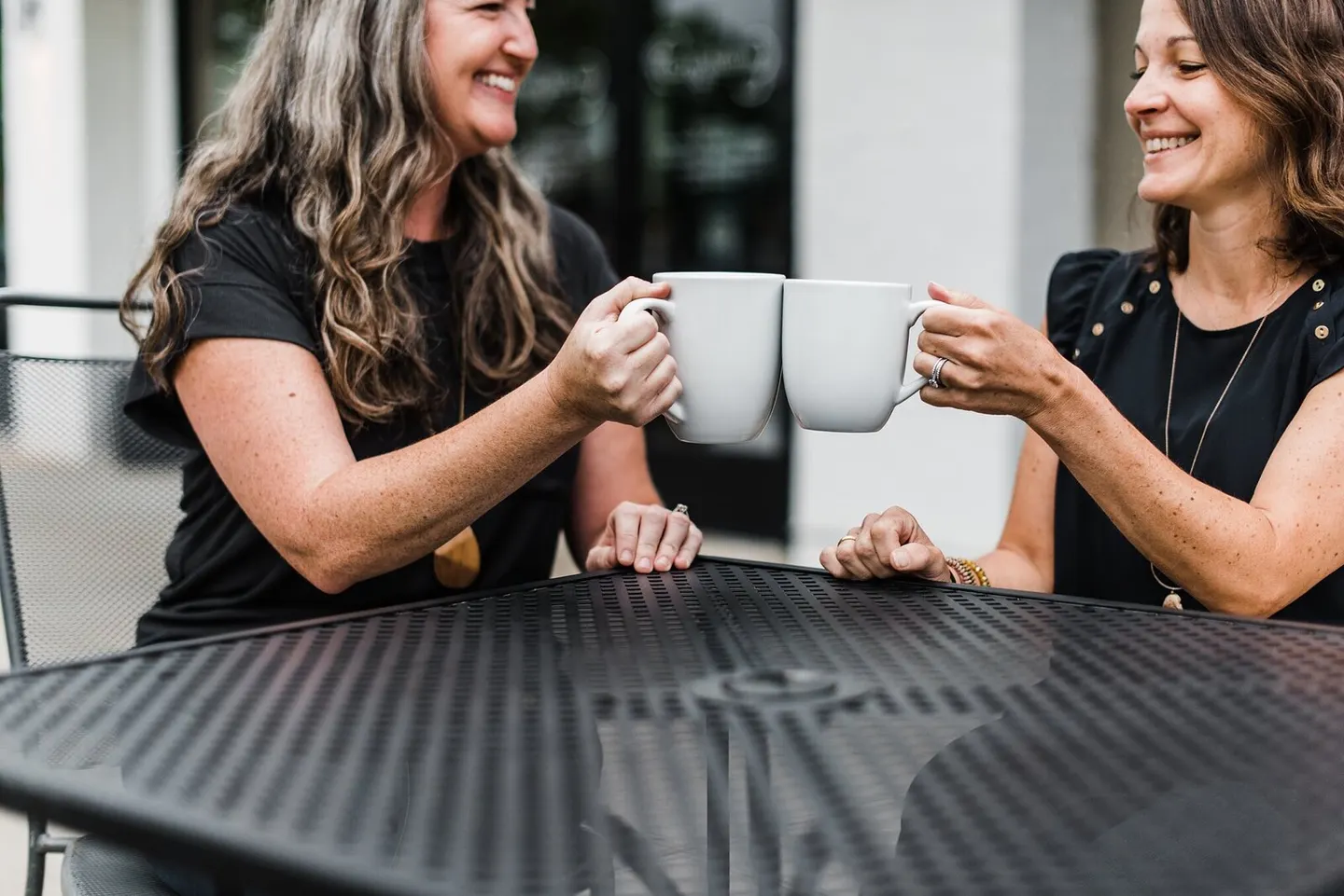 Two business partners share a laugh clinking coffee mugs at an outdoor cafe table