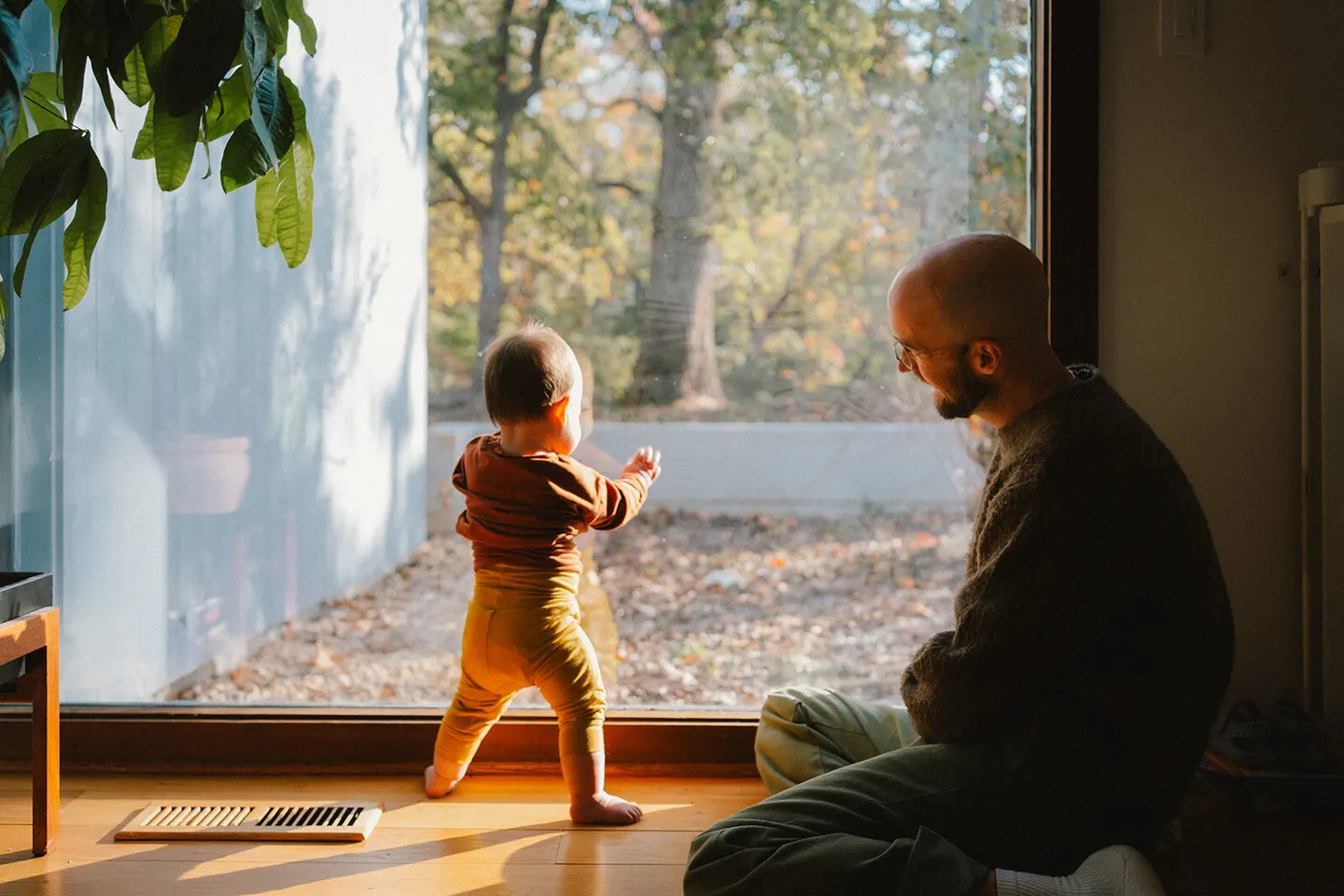 A father sits with his toddler at a floor-to-ceiling window, autumn trees glowing outside.