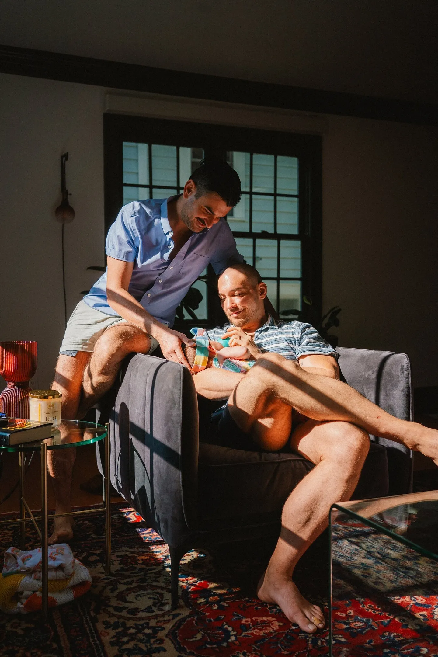 Two dads cradling their newborn on a sunlit armchair at home in Charlottesville