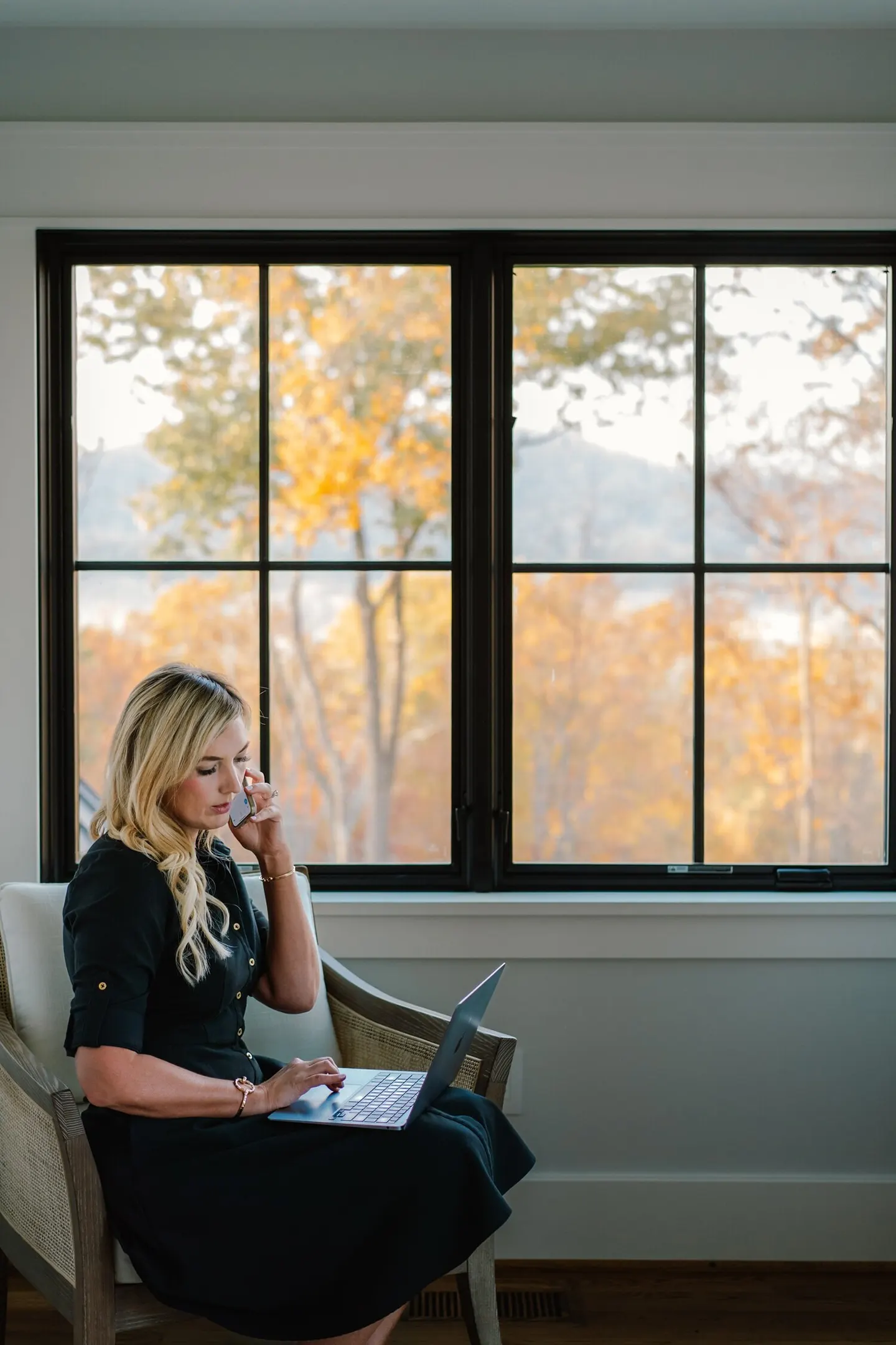 Real estate agent on the phone while typing on a laptop before a window framed by autumn trees