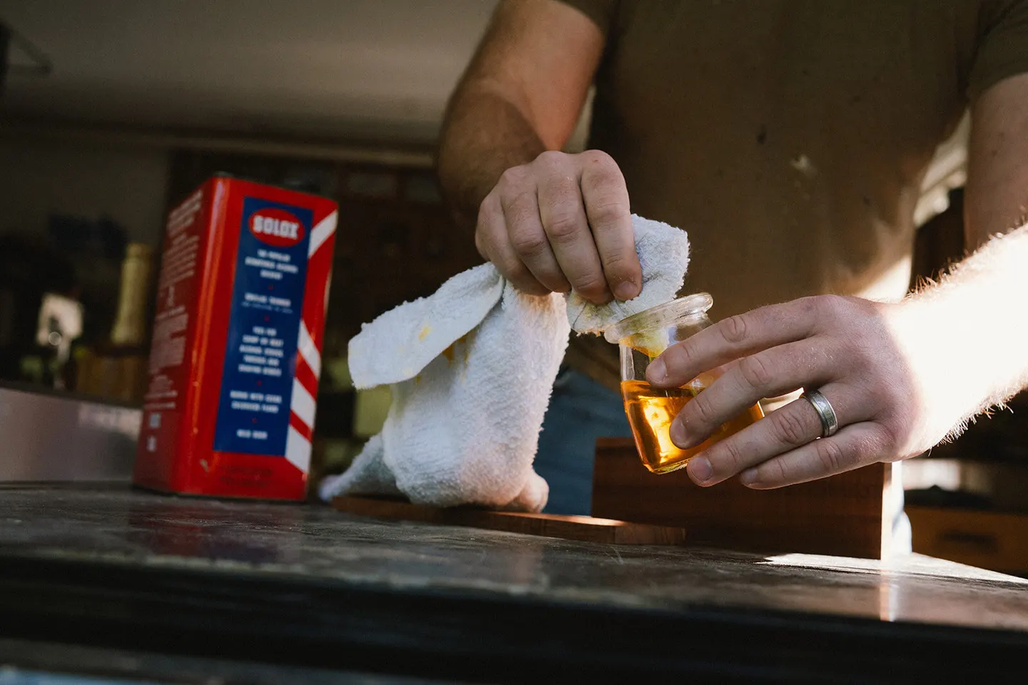 Woodworker's hands pour finishing oil into a small jar beside a vintage wax tin on a workbench
