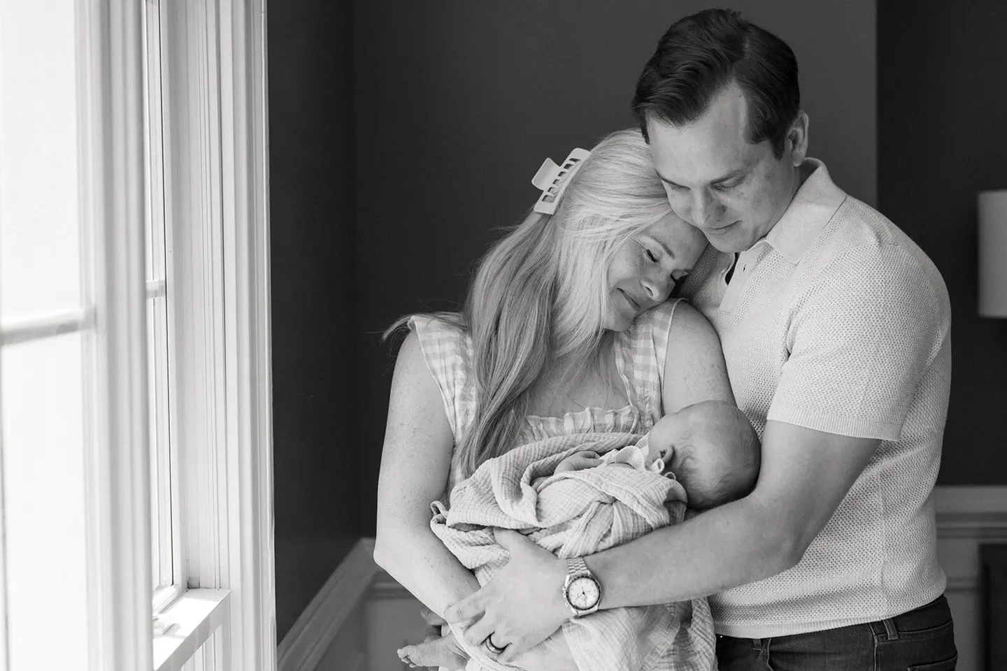 Parents huddle together cradling their swaddled newborn near a bright window