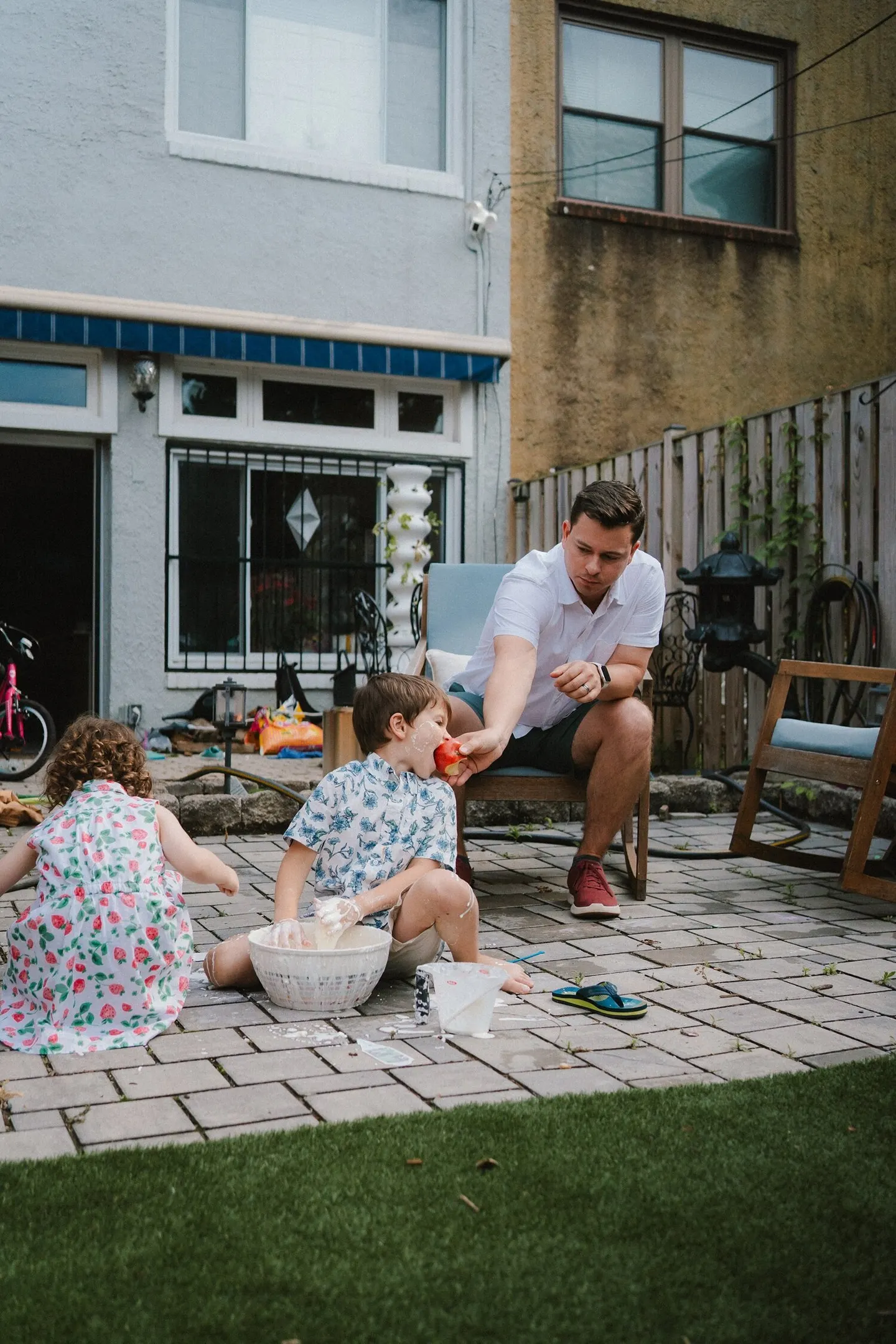On a backyard patio, a dad feeds his son a slice of apple while a girl snacks nearby.